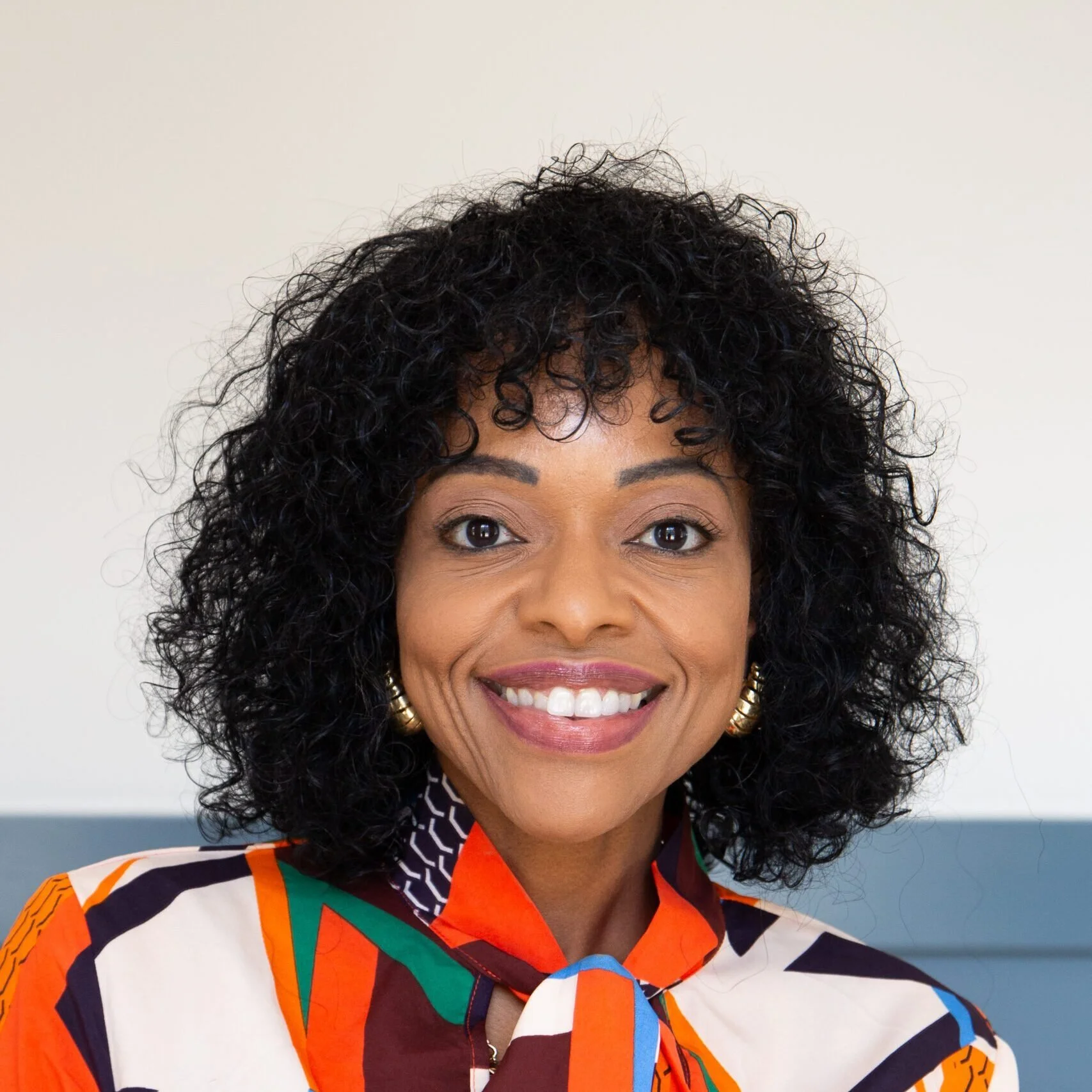 Close-up of woman with curly black hair, smiling, wearing large gold hoop earrings and a colorful patterned shirt, against a plain light background.