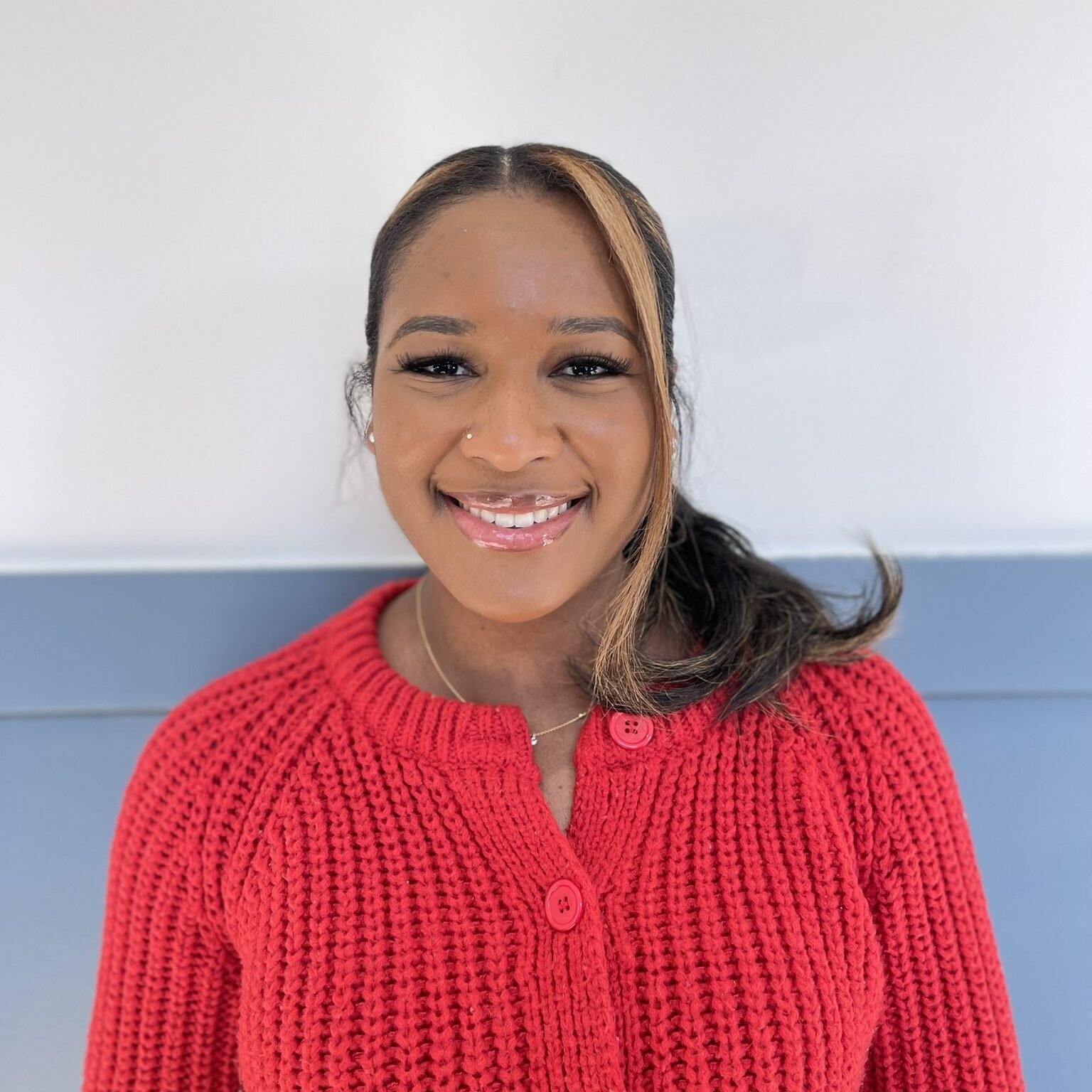 A smiling woman with medium skin tone, wearing a red knitted sweater and a small nose piercing, standing in front of a white and blue wall.