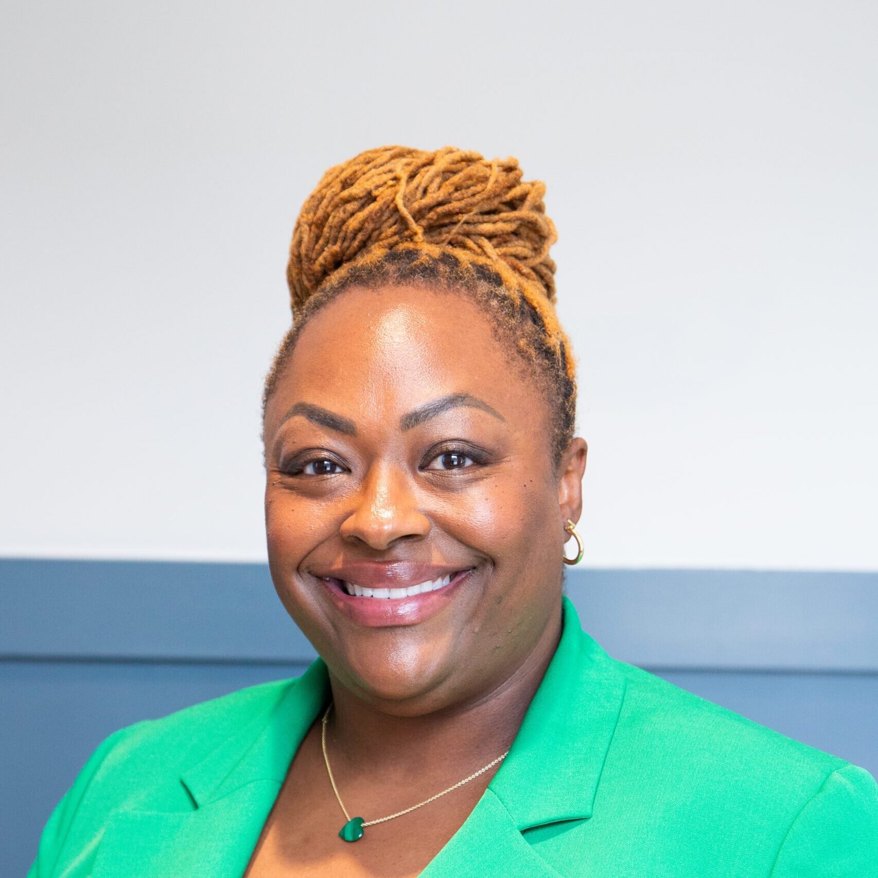 A smiling woman with short natural hair and a top knot styled with blonde dreadlocks, wearing a bright green blazer, hoop earrings, a heart-shaped pendant necklace, and a subtle makeup look, standing against a minimalist background.