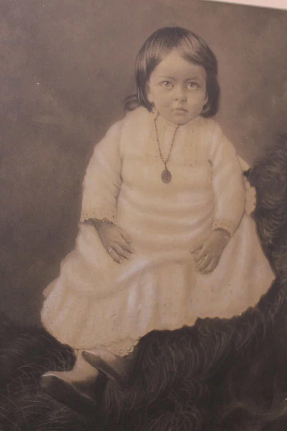 A vintage black and white photograph of a young girl with dark hair wearing a light-colored dress, a necklace, and a ring on her finger. She has a serious expression and is sitting with her hands on her lap.