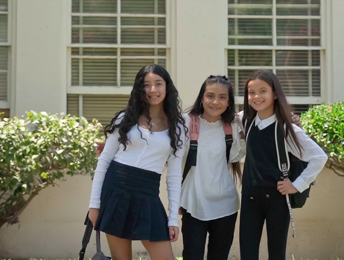Three young girls standing outdoors in front of a building with large windows, smiling, with backpacks on their shoulders.