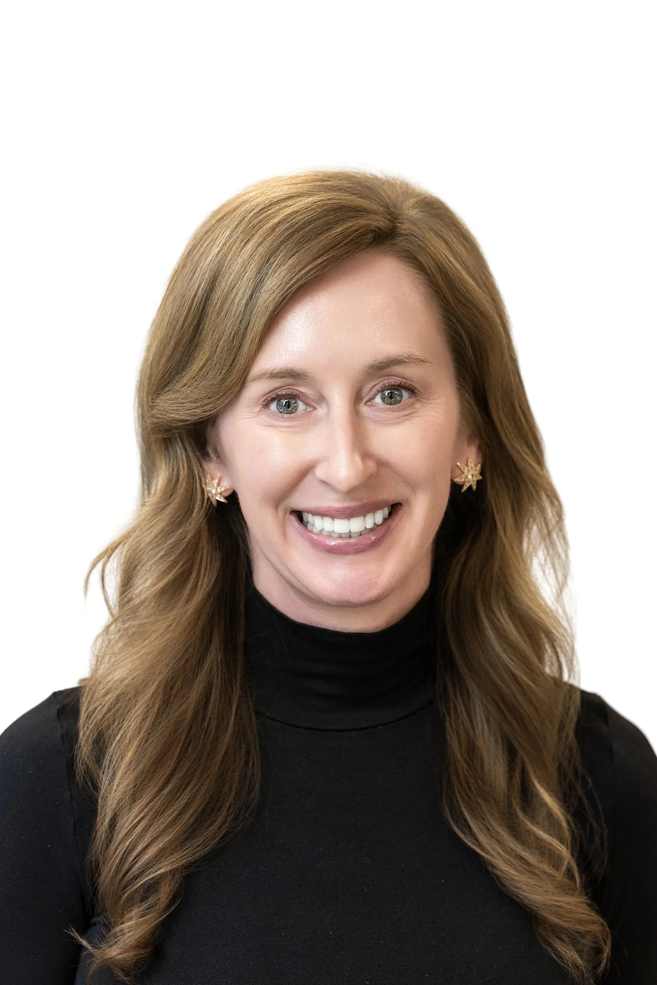 Close-up of a woman with light skin, brown hair, blue eyes, smiling, wearing gold floral earrings, blue top, against a plain light background.