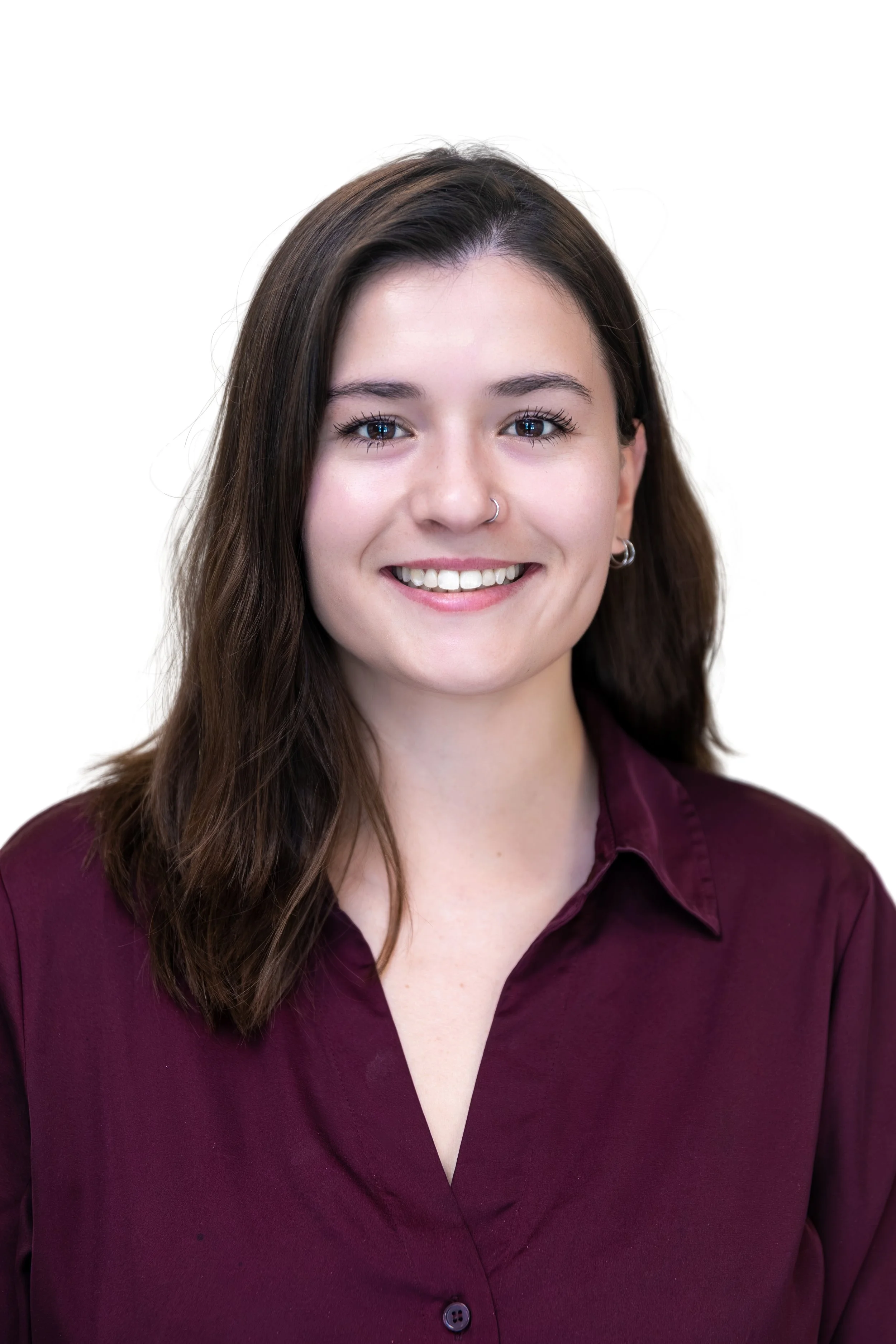 Close-up of a woman's face with brown hair, smiling, wearing a silver nose ring and earrings, against a neutral background.