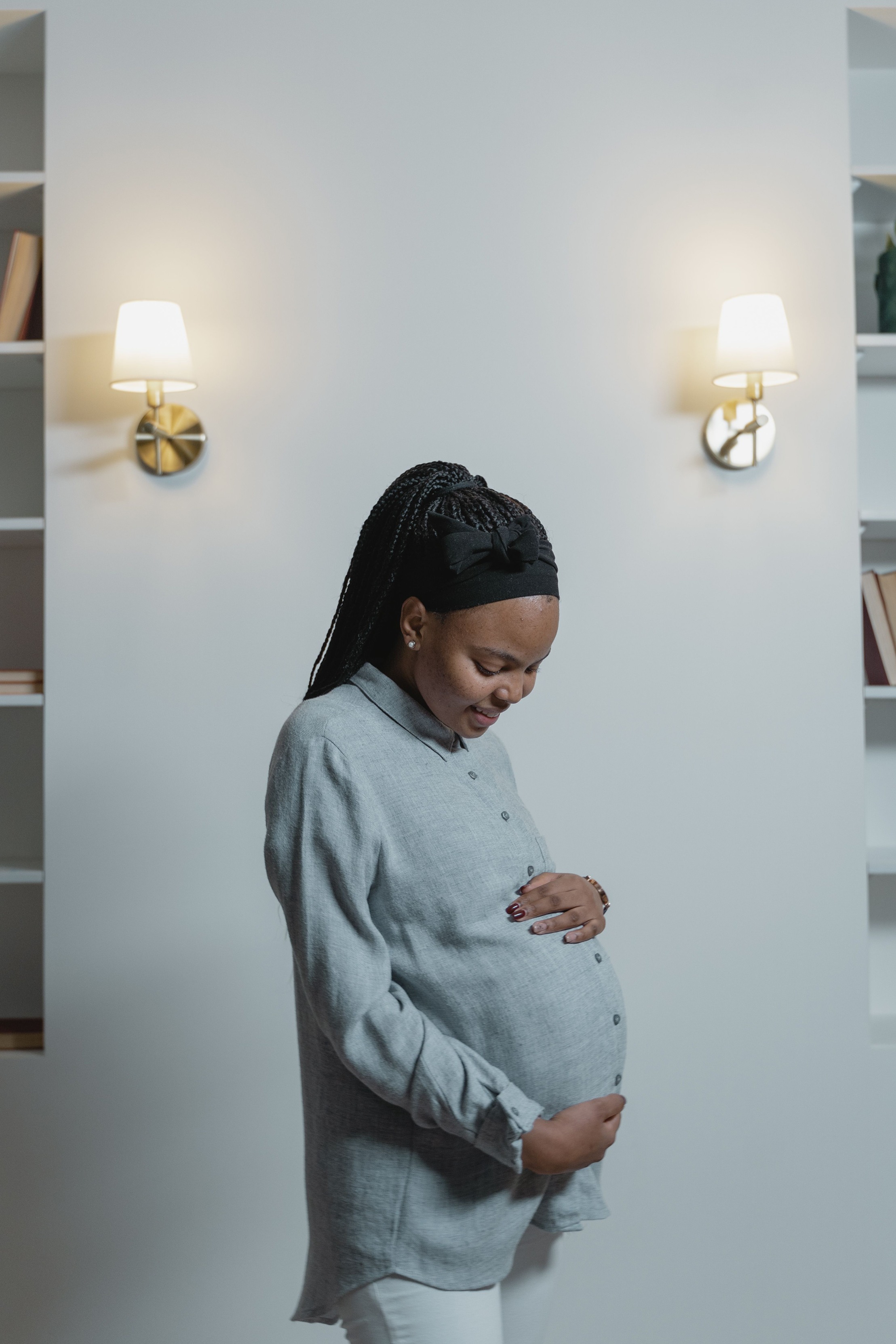 A pregnant woman with braided hair wearing a gray shirt and a black headband with a bow, smiling and looking down, standing in front of a white wall with two wall-mounted lamps.