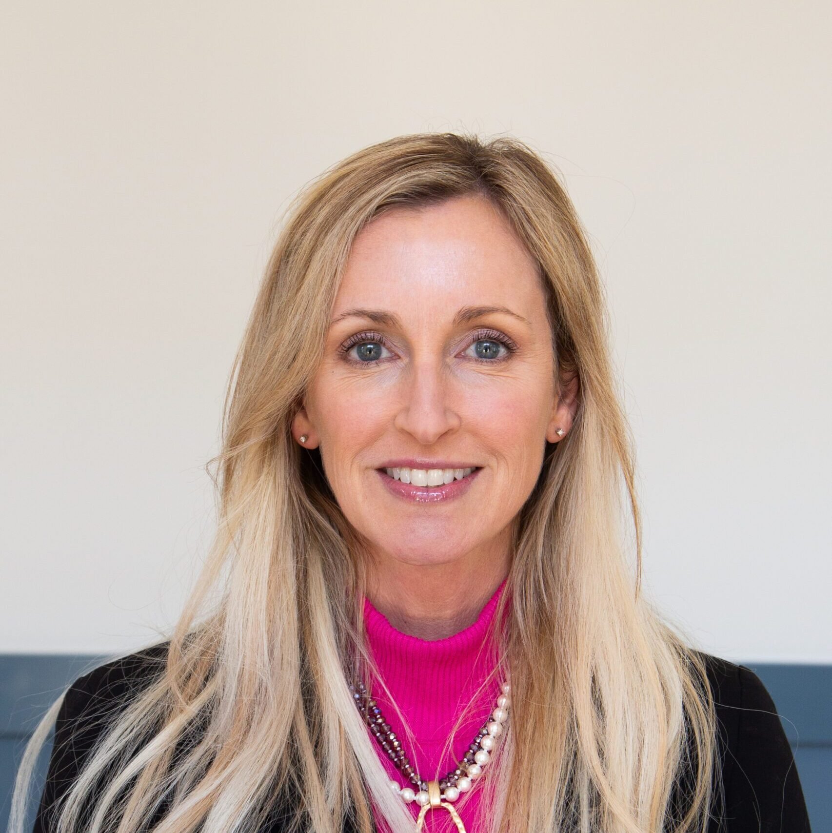 Close-up of a smiling woman with blonde hair, blue eyes, wearing a bright pink turtleneck, pearl earrings, and layered pearl necklaces, against a plain light background.