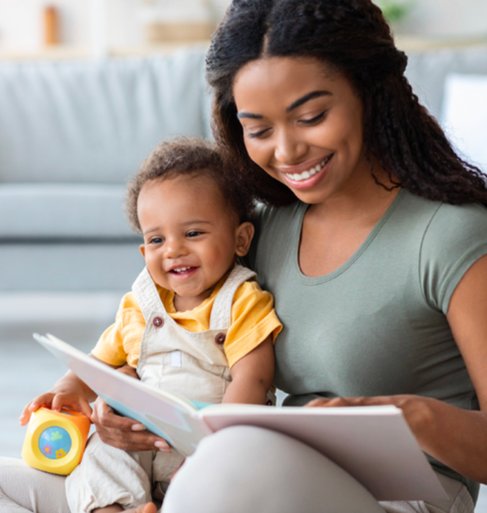 A woman and a young girl sitting on a light-colored couch, looking at a book together. The woman is smiling and the girl is happy, holding a colorful toy. The background shows a blurred living room with natural light.