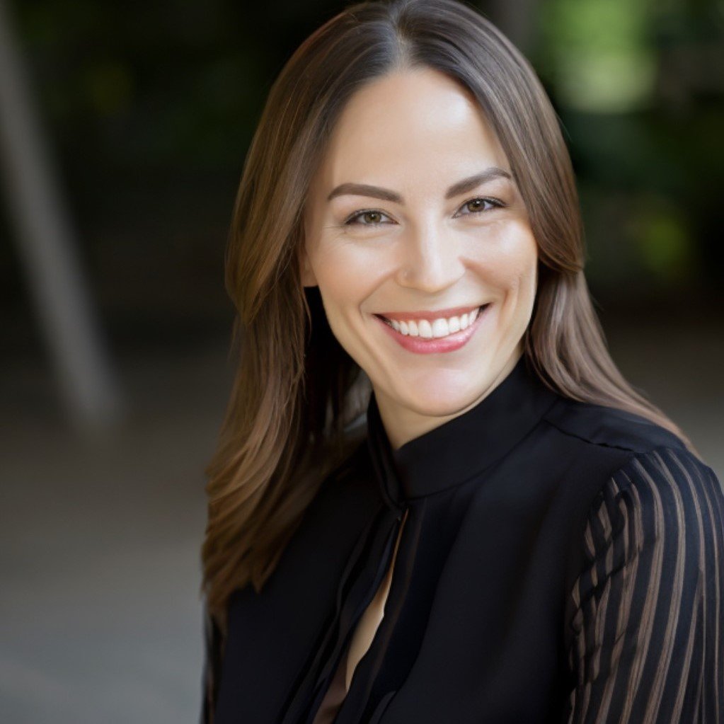 A smiling woman with light brown hair and fair skin, wearing a black top with sheer striped sleeves, outdoors with blurred greenery in the background.