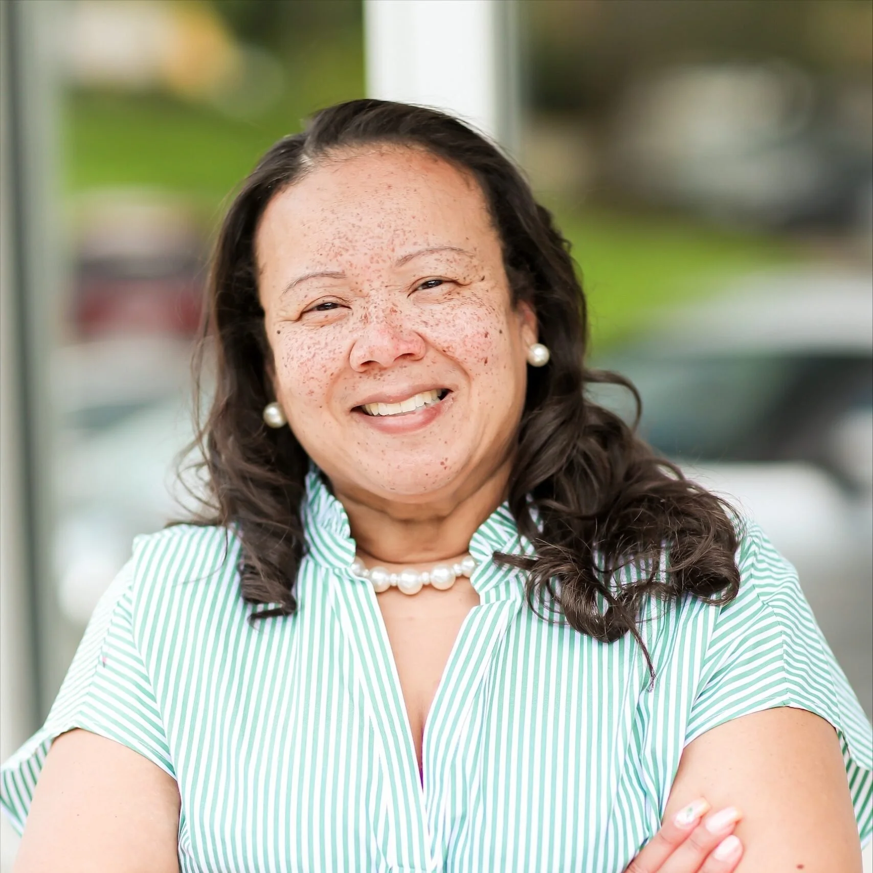 A woman with dark wavy hair, wearing pearl earrings and a green and white striped shirt, smiling with her arms crossed in front of a blurred outdoor background.
