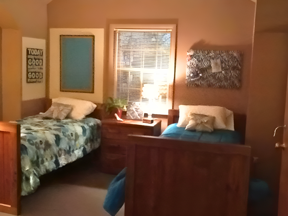 A cozy bedroom with two beds, one with a floral bedspread and the other with a blue bedspread, sharing a wooden nightstand with a lamp and plants. There are pillows, wall art, a window with blinds and a lamp, and warm lighting.