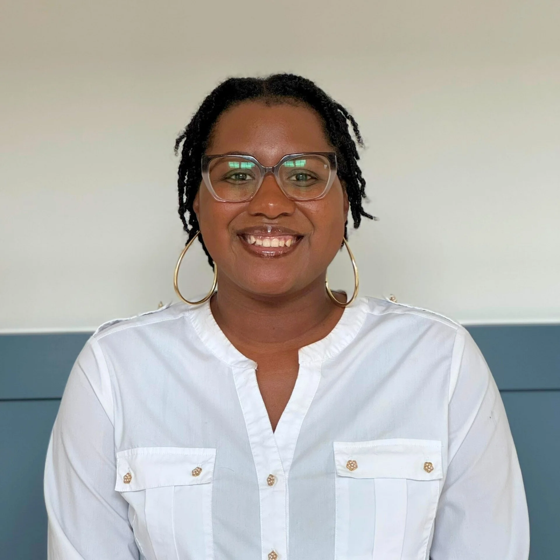 A woman smiling, wearing glasses, gold hoop earrings, and a white blouse with decorative gold buttons, standing against a plain light-colored wall.