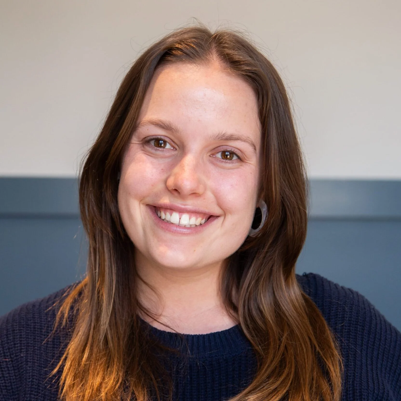 A woman with long brown hair, smiling, wearing a navy blue sweater, against a plain background.