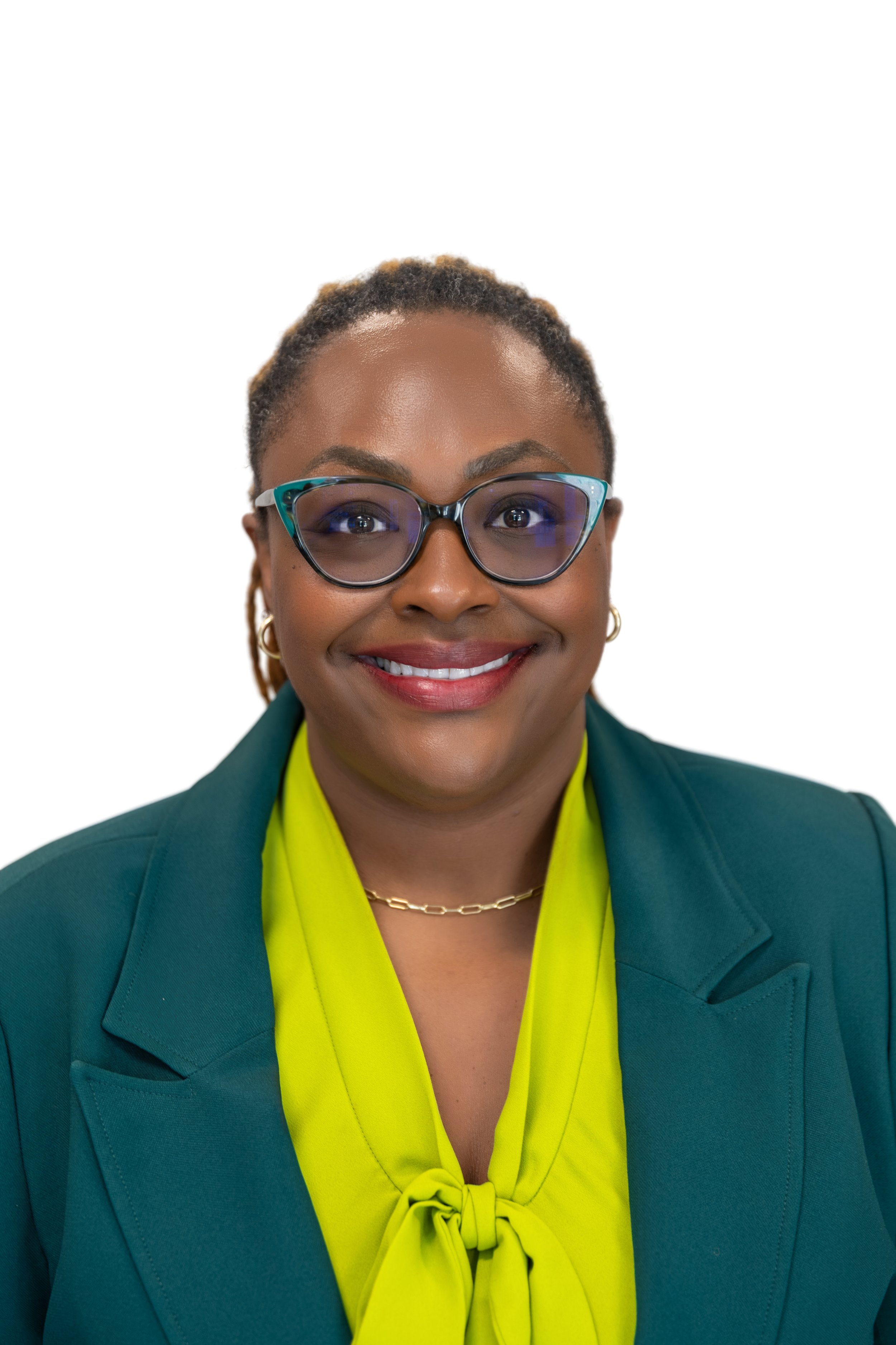 A smiling woman with short natural hair and a top knot styled with blonde dreadlocks, wearing a bright green blazer, hoop earrings, a heart-shaped pendant necklace, and a subtle makeup look, standing against a minimalist background.