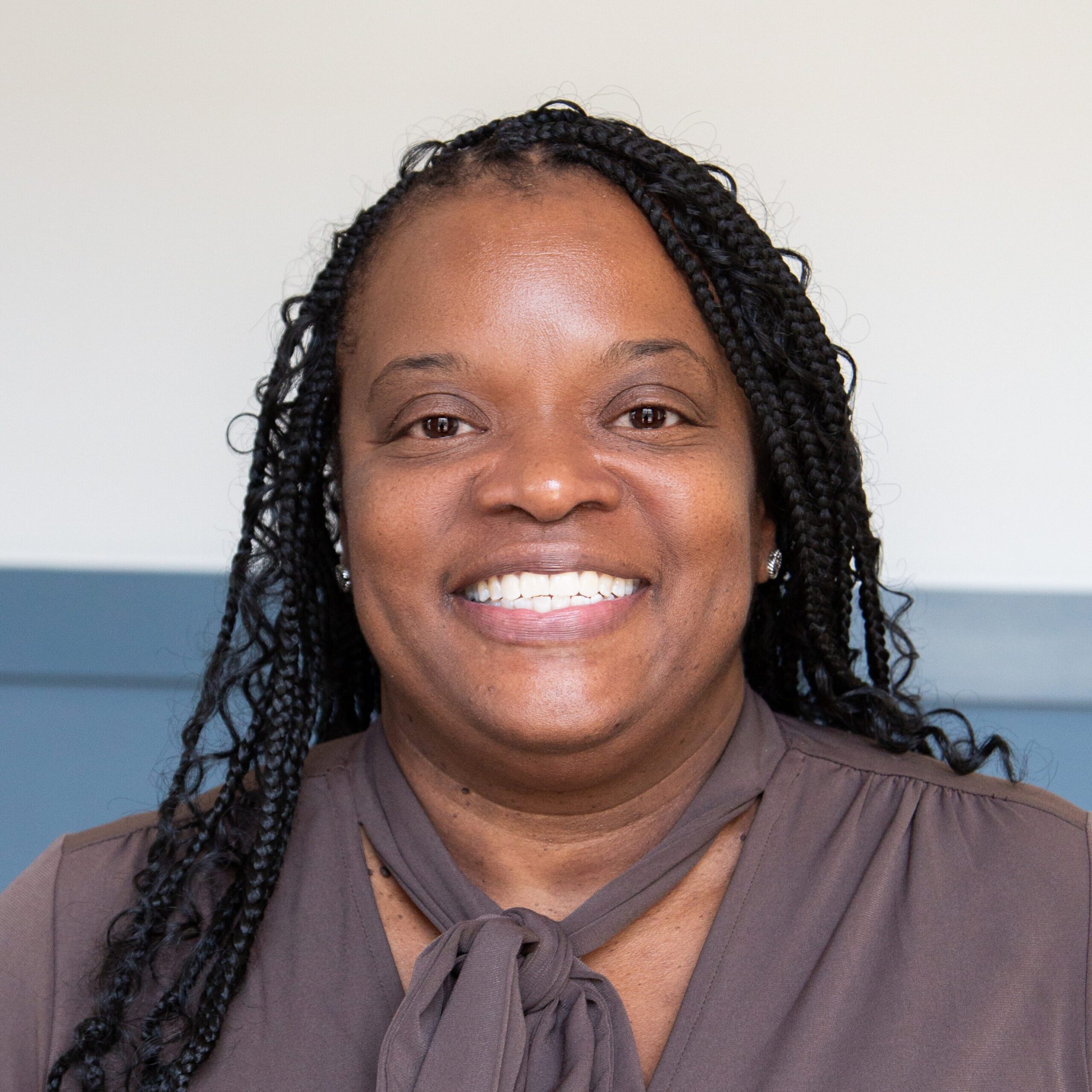 Close-up of a woman with dark braided hair, smiling, wearing a brown top and a matching brown tie-front shirt.