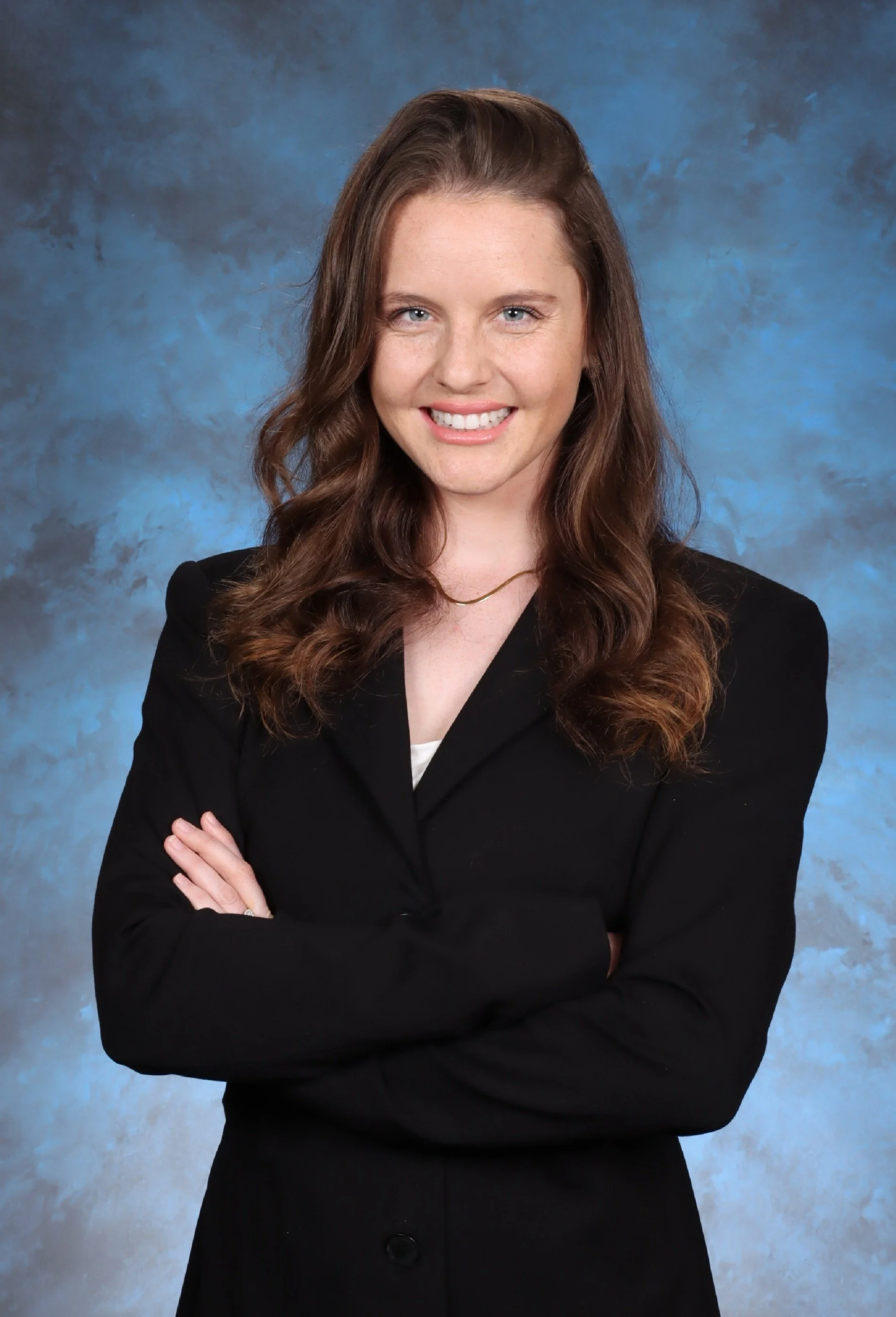 A female attorney with mediation skill, with long brown hair, wearing a black blazer, cross arms, smiling, against a blue mottled background.