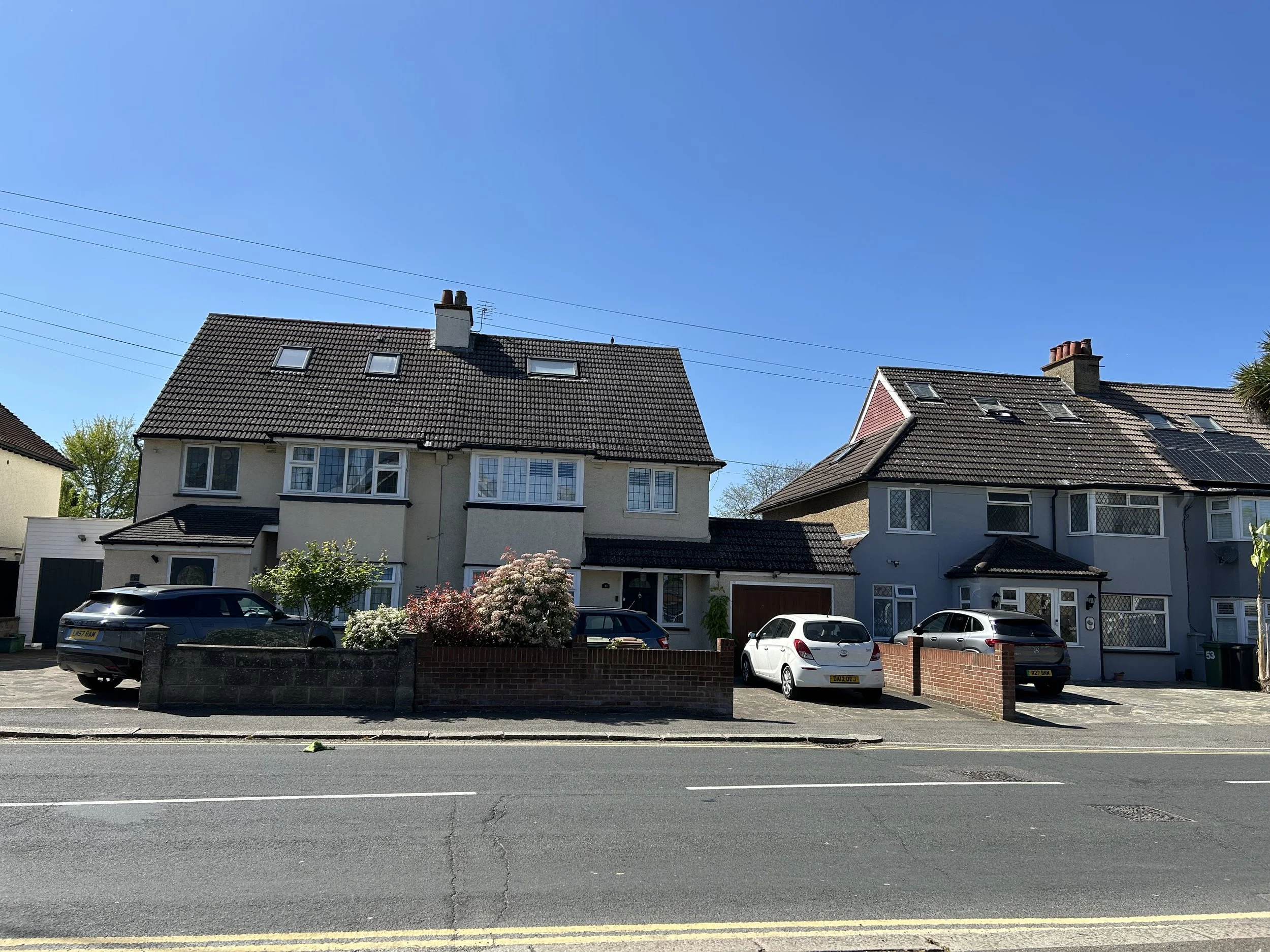 A residential street with two houses, parked cars, trees, and a clear blue sky.