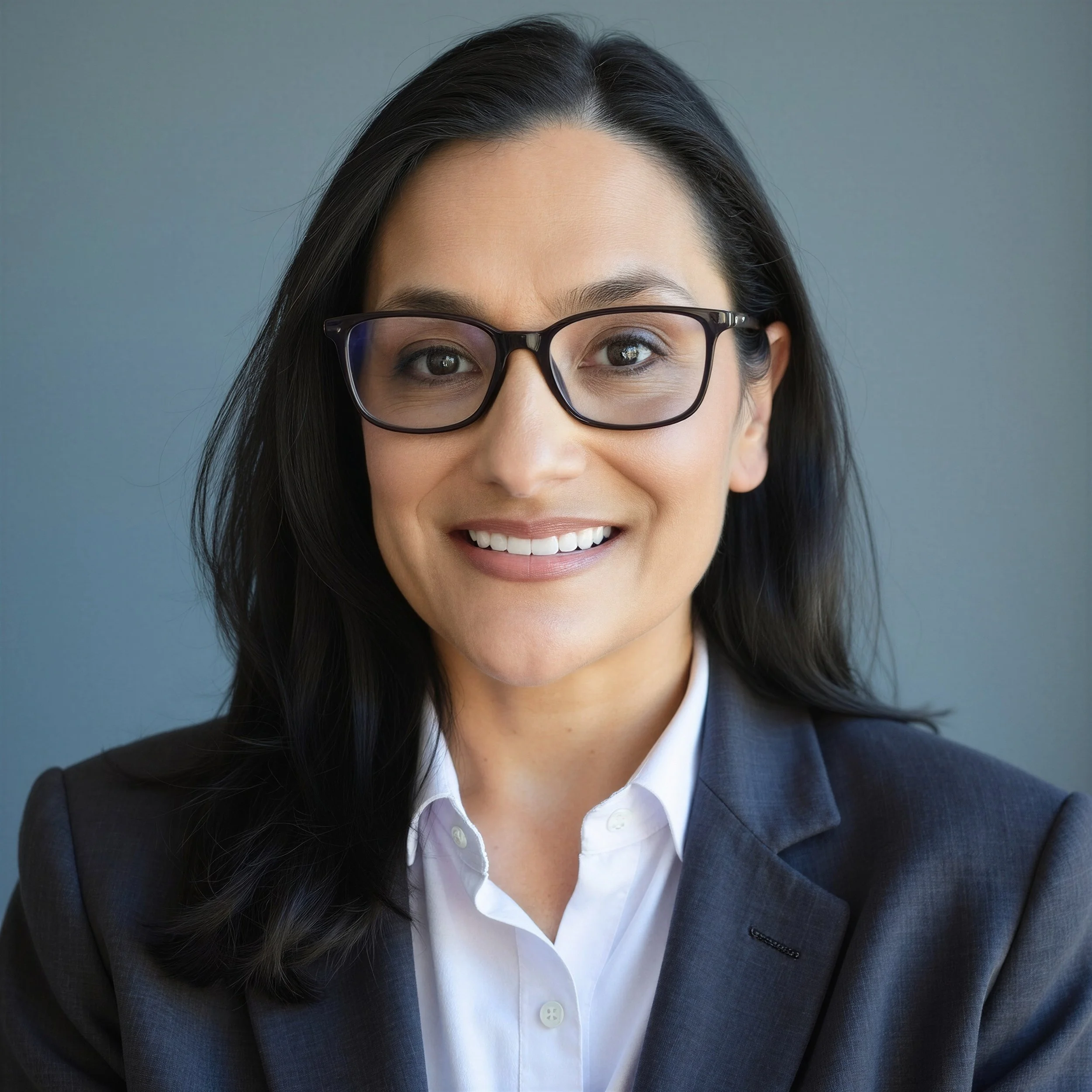 Close-up of a smiling woman with black hair, wearing glasses, a white shirt, and a dark blazer, against a gray background.