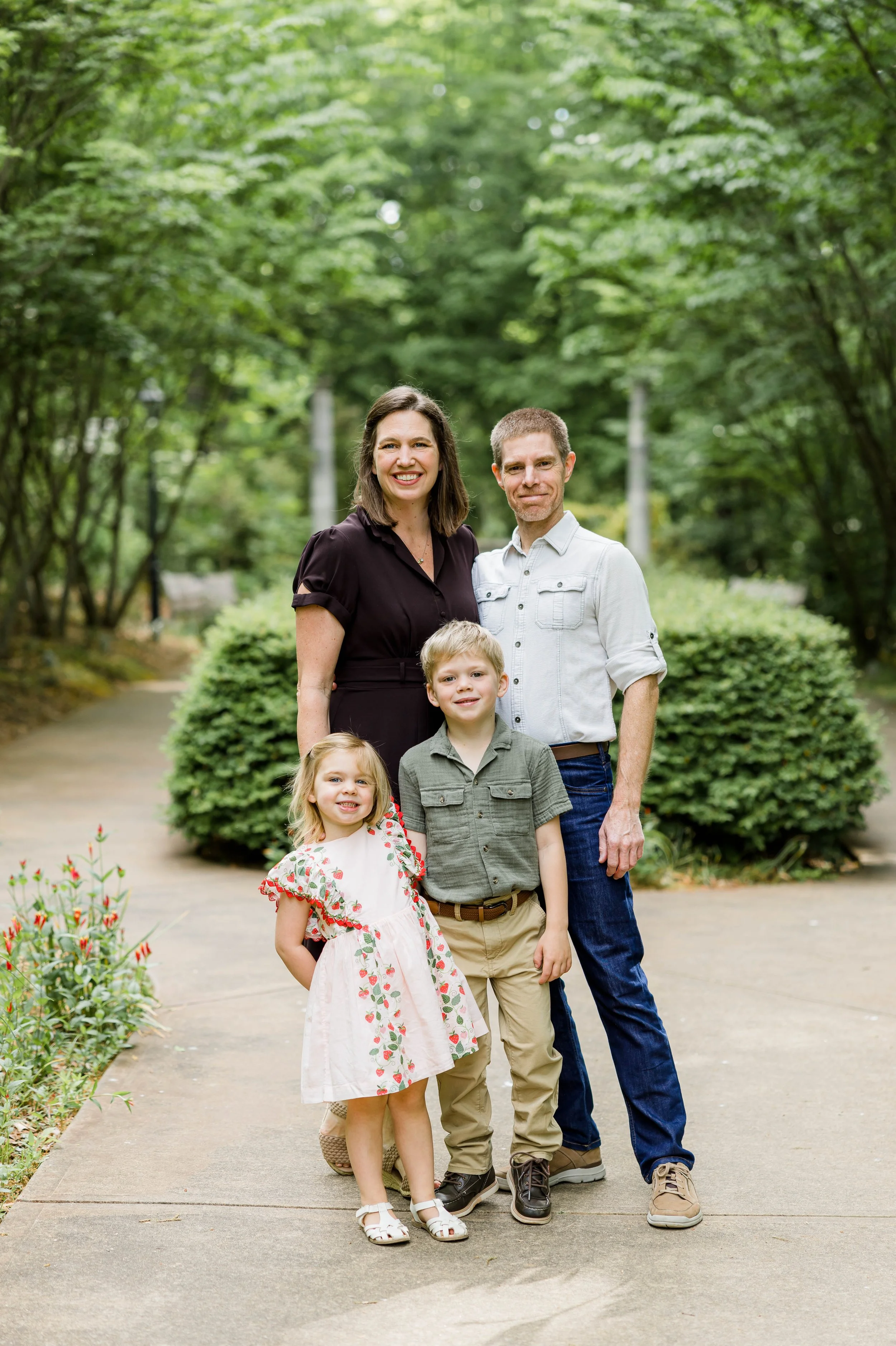 A family of five standing on a sidewalk in a lush green park, smiling at the camera. The group includes a mother, father, and three young children, with trees and bushes in the background.