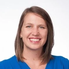 A woman with shoulder-length brown hair, smiling, wearing a blue top, against a white background.