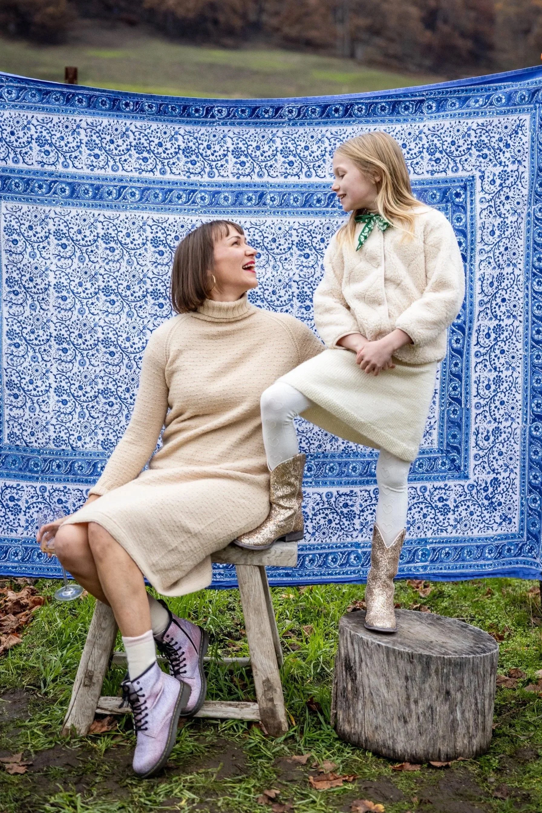 A woman and a girl having fun outdoors, with a blue patterned blanket hanging behind them. The woman is seated on a small wooden stool, wearing a beige dress and boots, smiling at the girl. The girl is standing on a tree stump, dressed in a cream-colored coat and white leggings, looking at the woman.