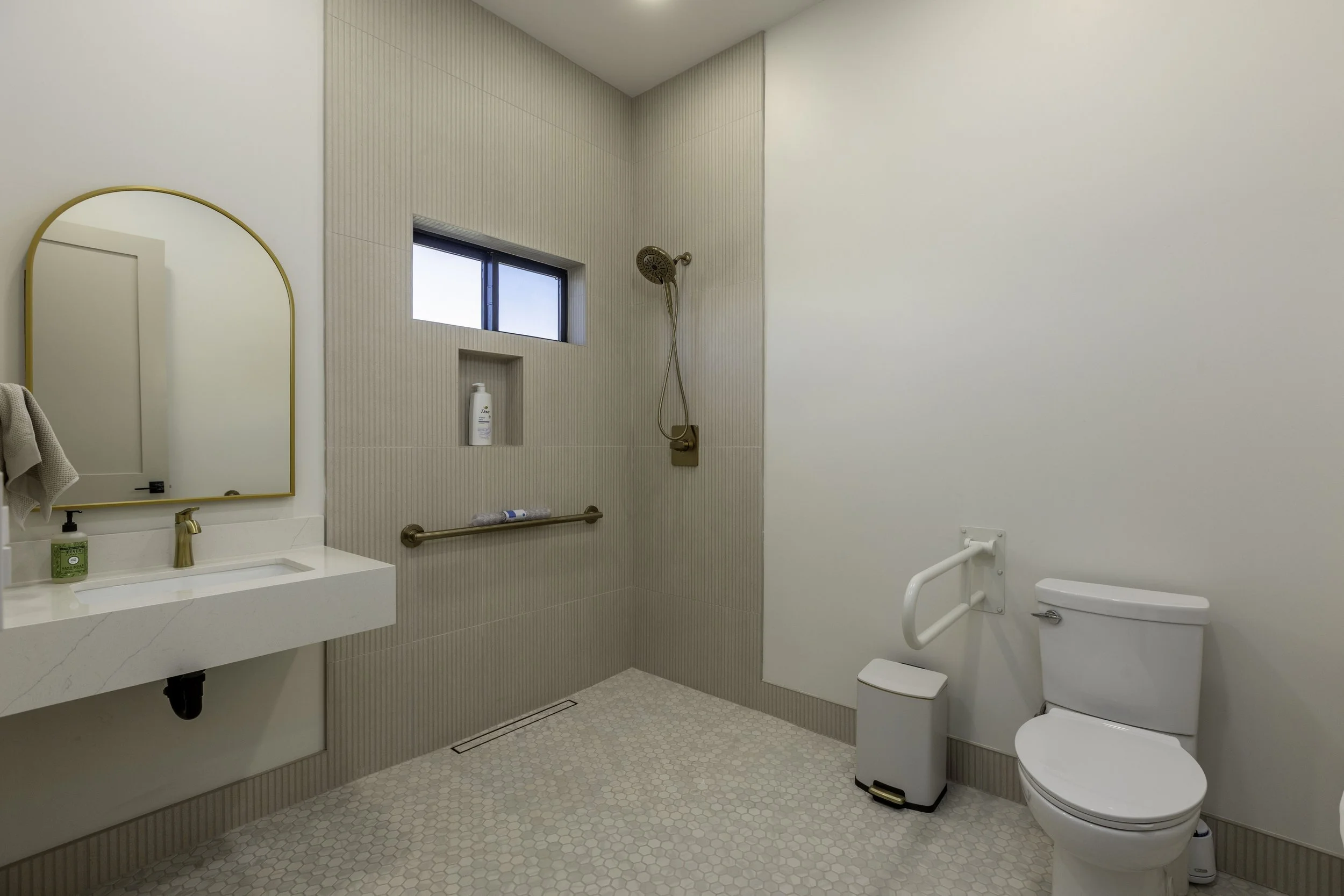 A modern bathroom with a white sink, gold faucet, mirror with gold frame, small window, showerhead, and toilet, featuring beige wall tiles and patterned floor tiles.
