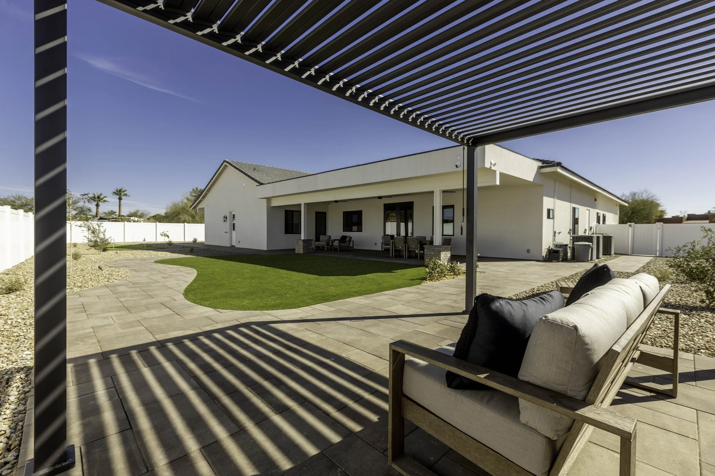 Back patio area view of a modern white house with patio furniture, covered by a metal pergola, with manicured lawn and landscaped yard, under a clear blue sky.