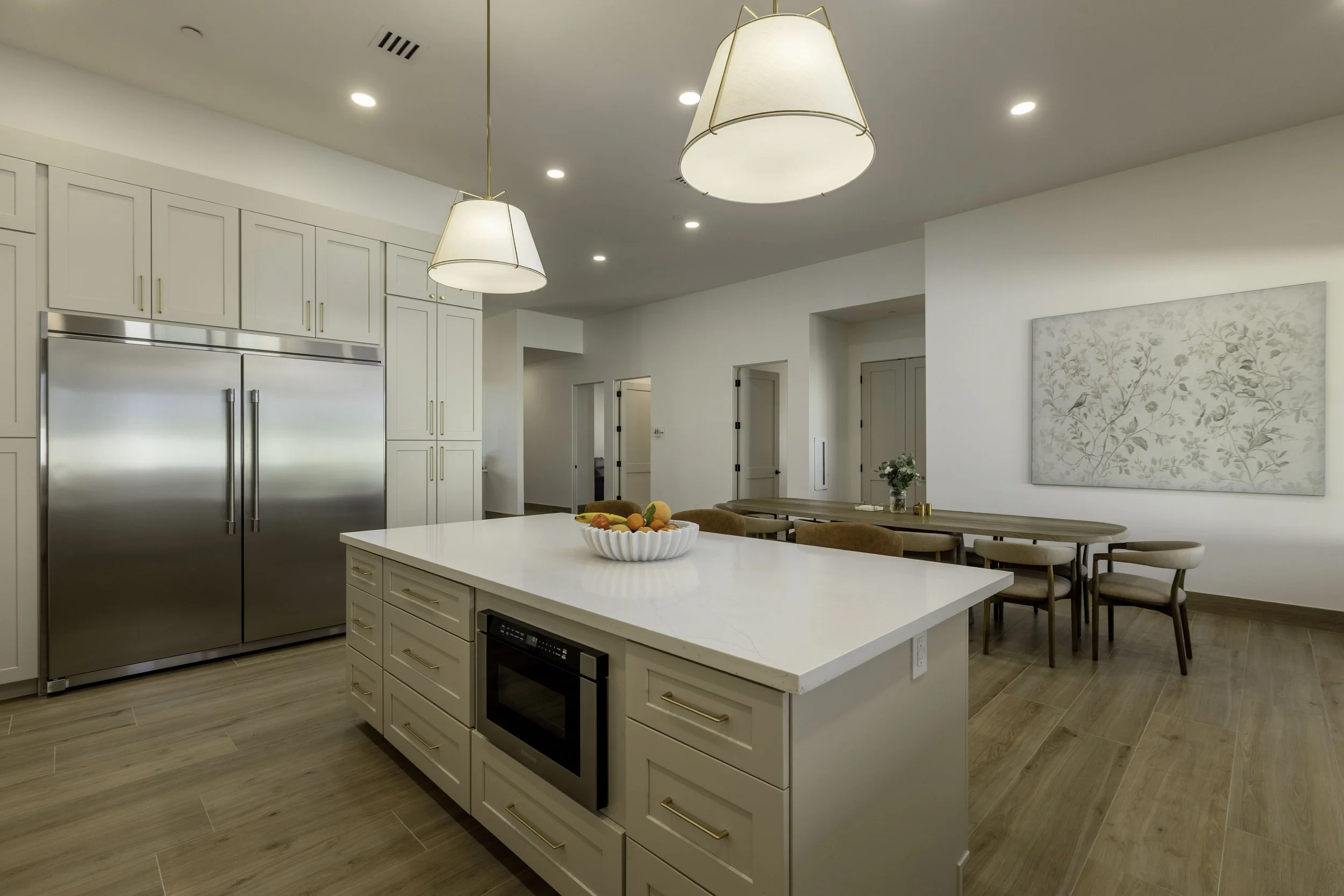 Modern kitchen with white cabinets, stainless steel refrigerator, white island with a bowl of fruit, wooden dining table with beige chairs, large pendant lights, and framed artwork on the wall.
