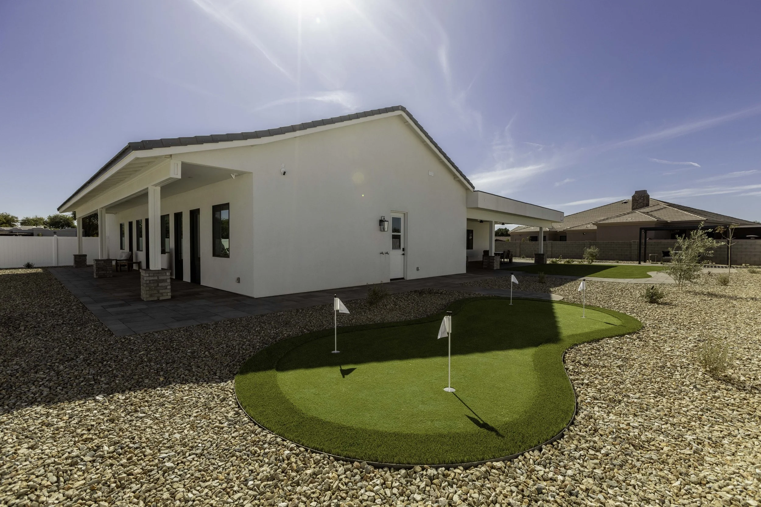Backyard with a putting green, gravel, and small trees, adjacent to a white house with a covered patio, under a clear blue sky.