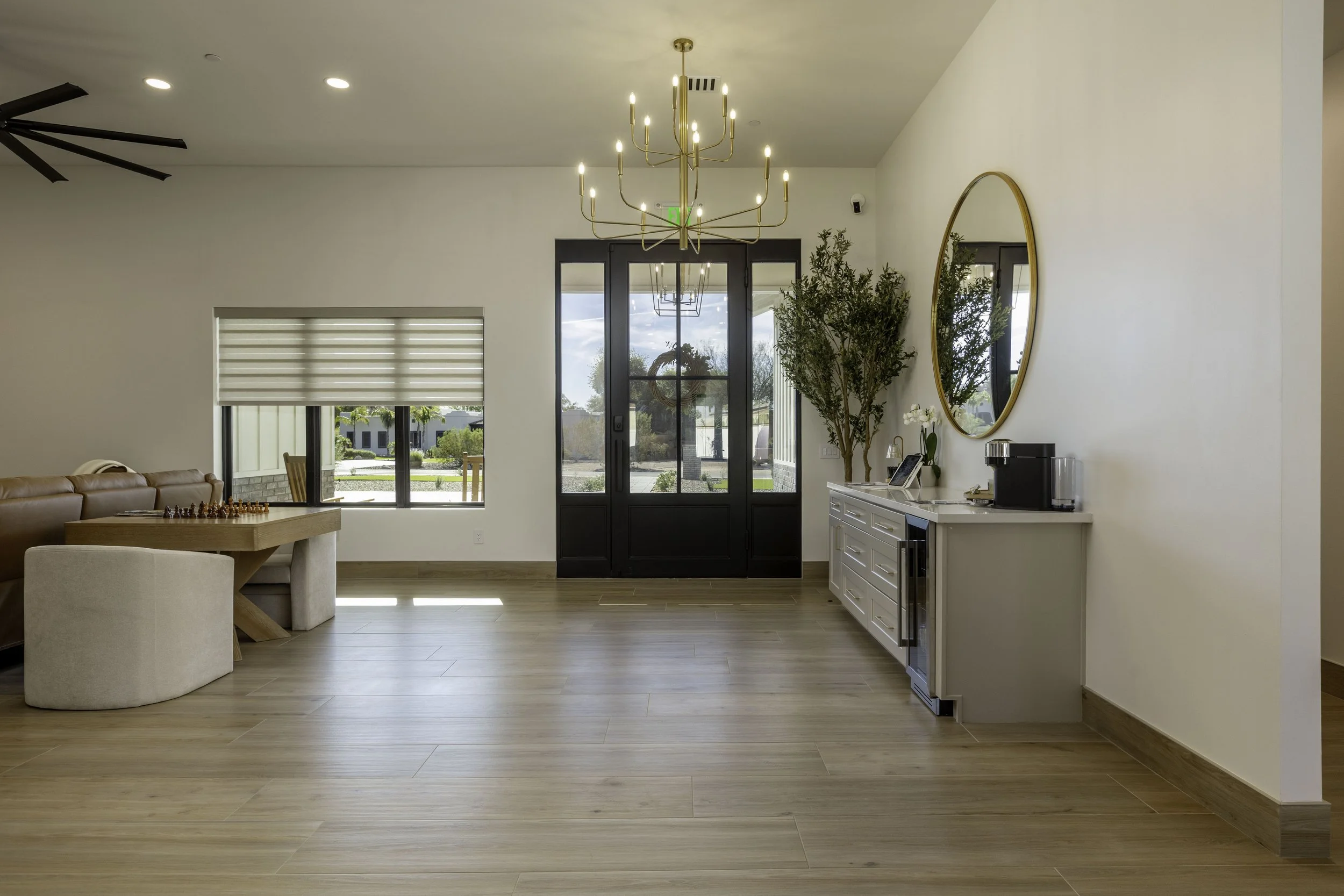 Interior view of a modern living space with a chandelier, large windows, a beige sofa, a wooden dining table with chairs, and a white sideboard with plants and a mirror.