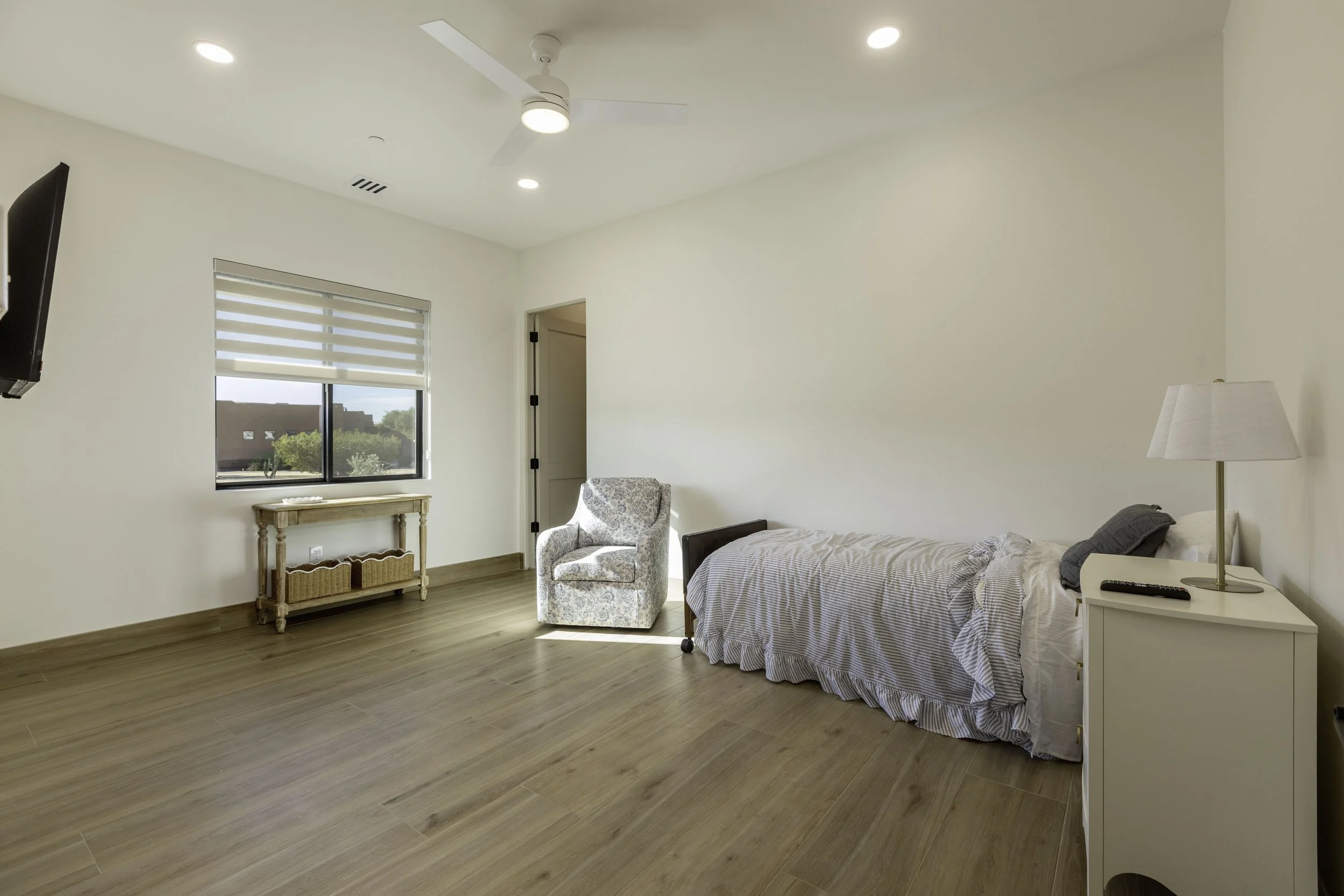 A bedroom with a bed, a nightstand with a lamp, a patterned armchair, a window with horizontal blinds, and a ceiling fan.