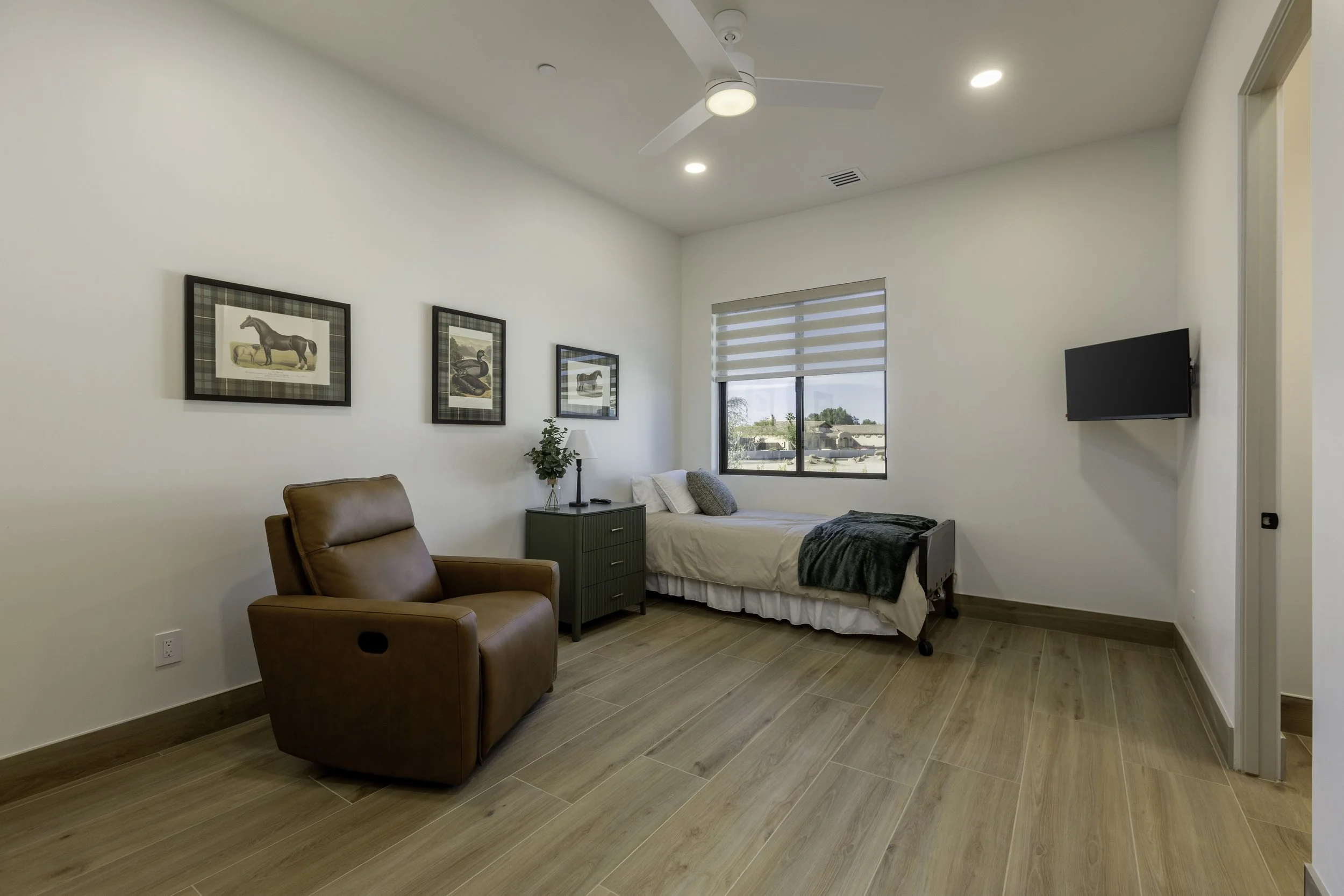 A bedroom with a single bed, a green nightstand, a brown leather armchair, and framed horse pictures on the wall. There is a window with blinds and a wall-mounted TV. The room has wooden flooring and a white ceiling with a ceiling fan and recessed lighting.