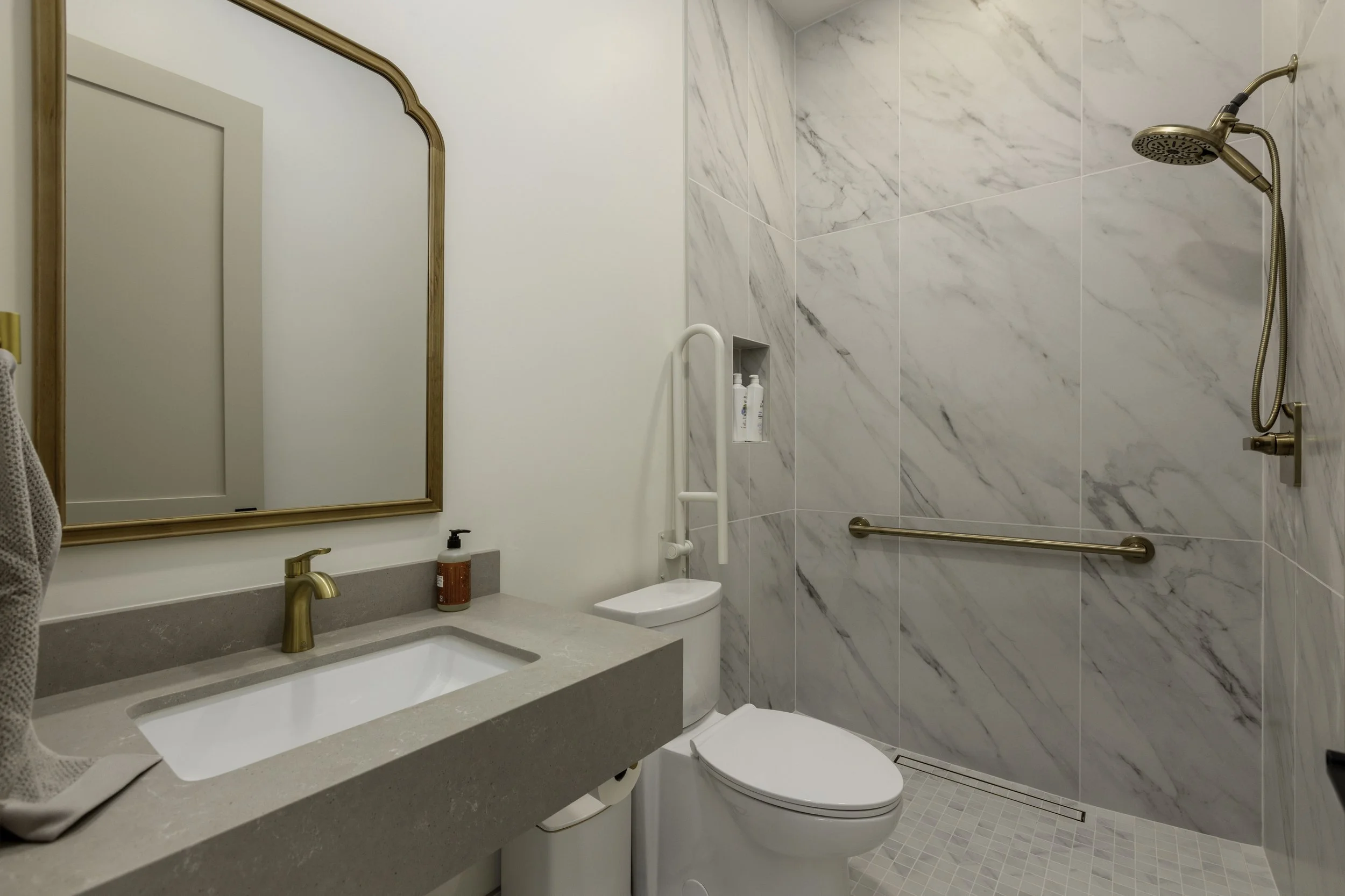 Modern bathroom with marble tile wall, white toilet, beige countertop, gold faucet, large mirror, and walk-in shower with grab bars and built-in shelves.