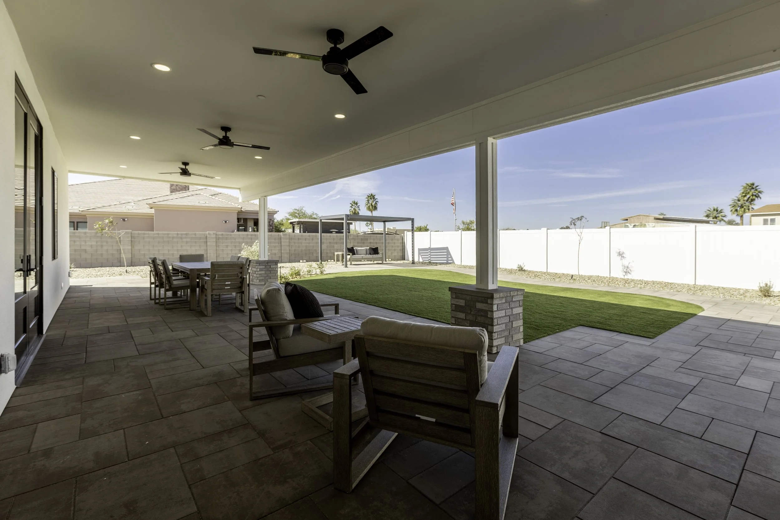 Covered backyard patio with outdoor seating, a grassy area, and a view of neighboring houses and palm trees under a clear blue sky.