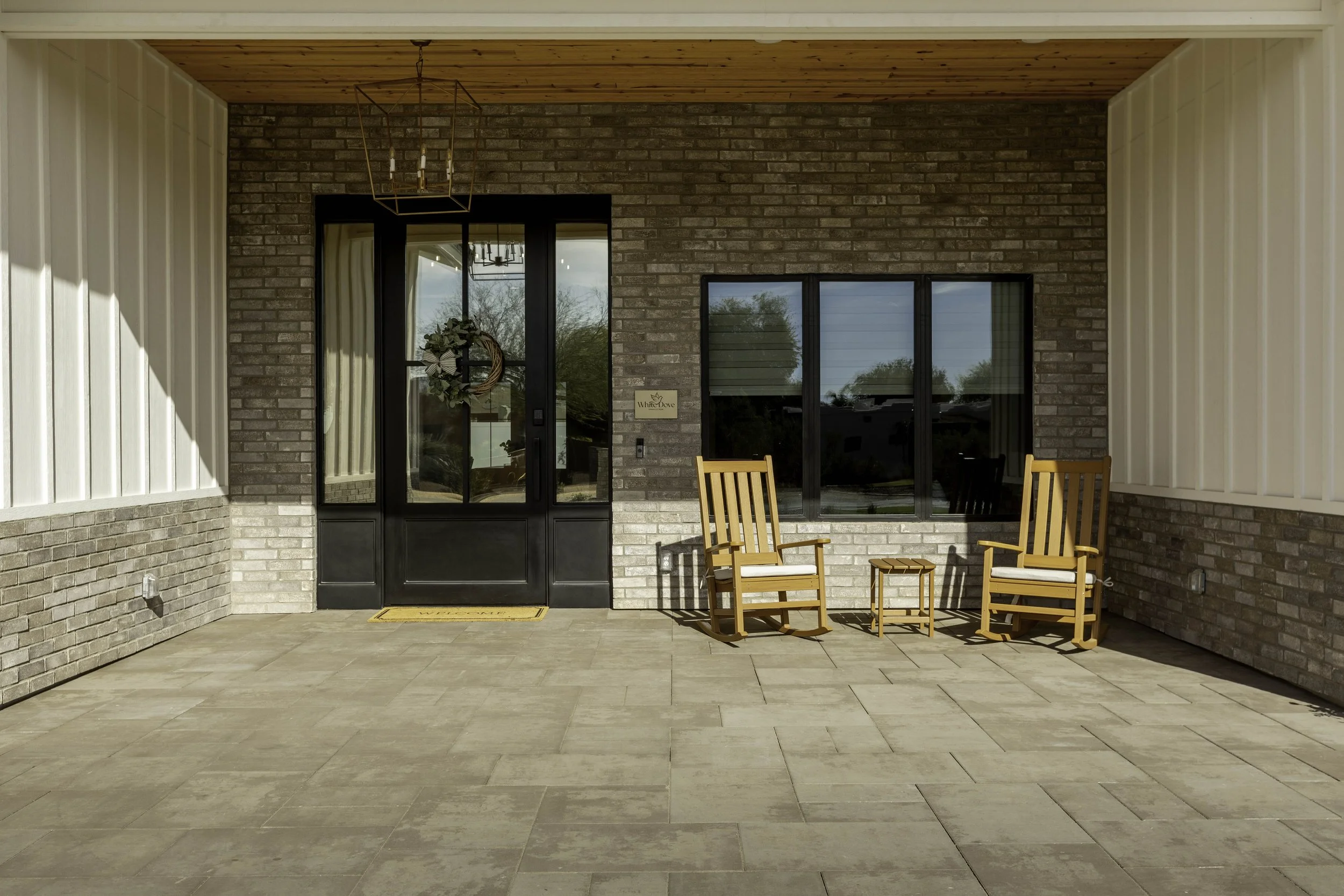 Covered patio with brick walls and a wood ceiling, featuring two wooden rocking chairs and a small table, with a door and window reflecting trees outside.