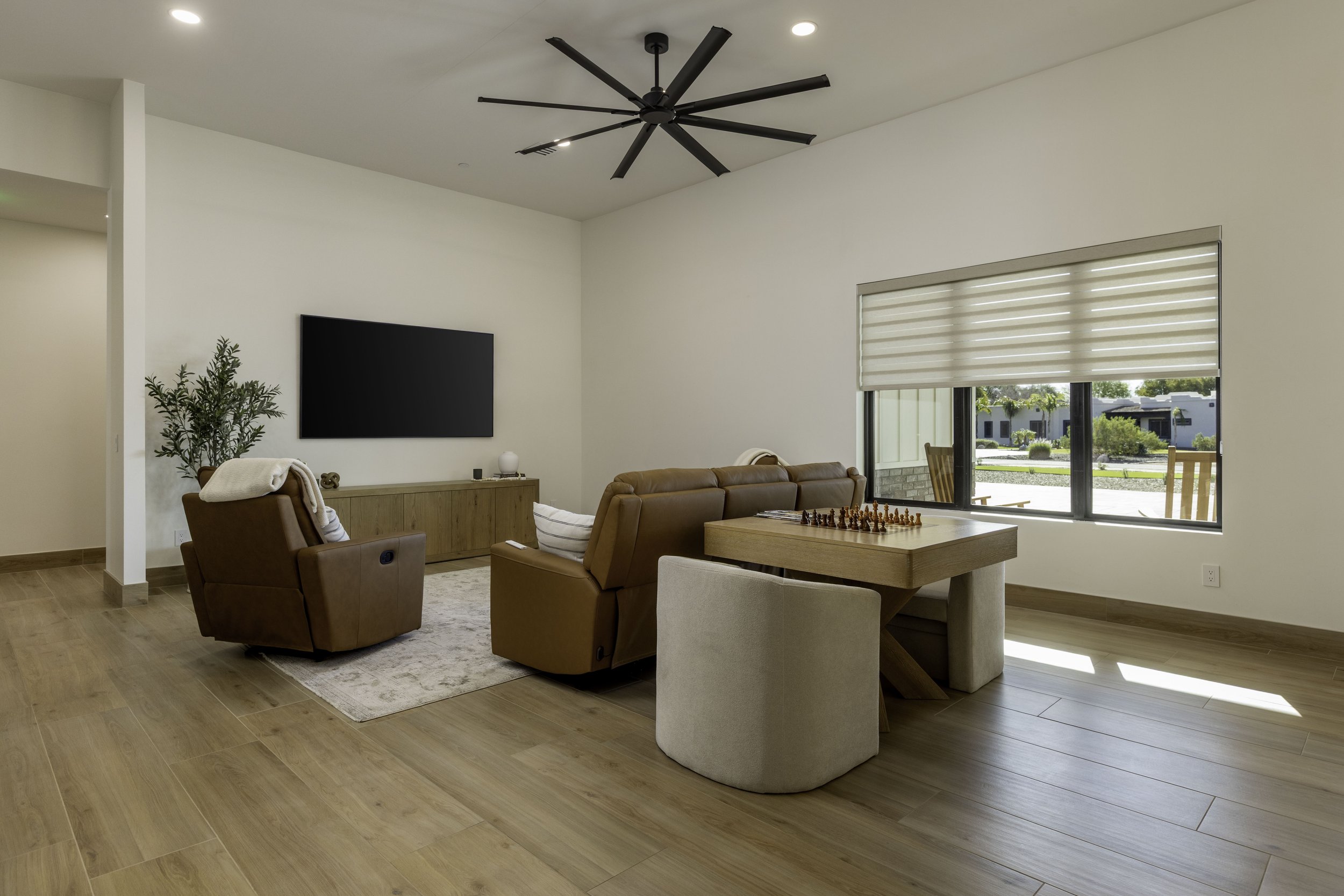 Living room with wooden flooring, tan leather chairs, a white upholstered chair, a wooden coffee table with a chessboard, a wall-mounted TV, a TV stand with decorative items, a large window with blinds, and a ceiling fan.