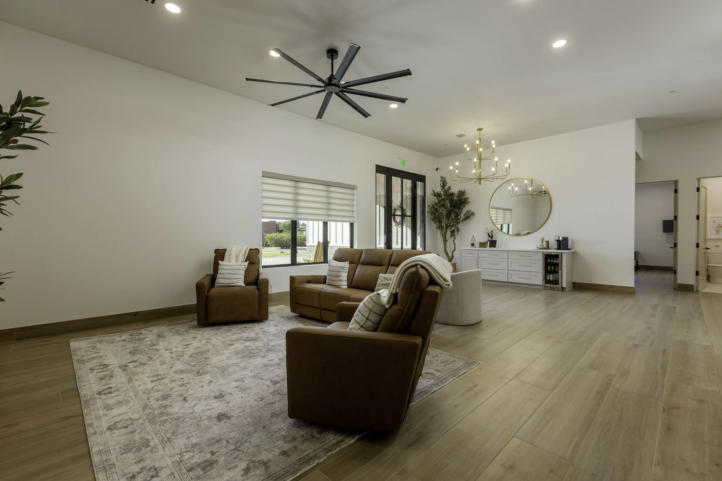 Modern living room with beige sofas, patterned rug, large window with blinds, decorative plants, a white sideboard, a round mirror, chandelier, and ceiling fans.