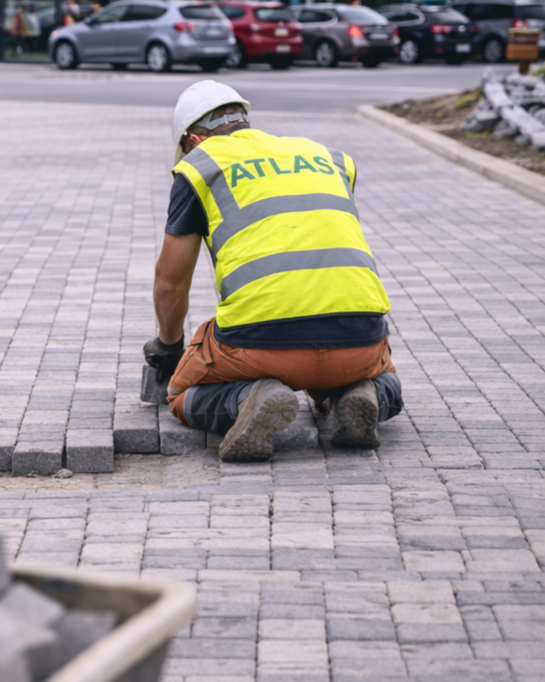 A worker wearing a yellow safety vest with Atlas Paving on the back and a white helmet, kneeling on block paving, installing paving.