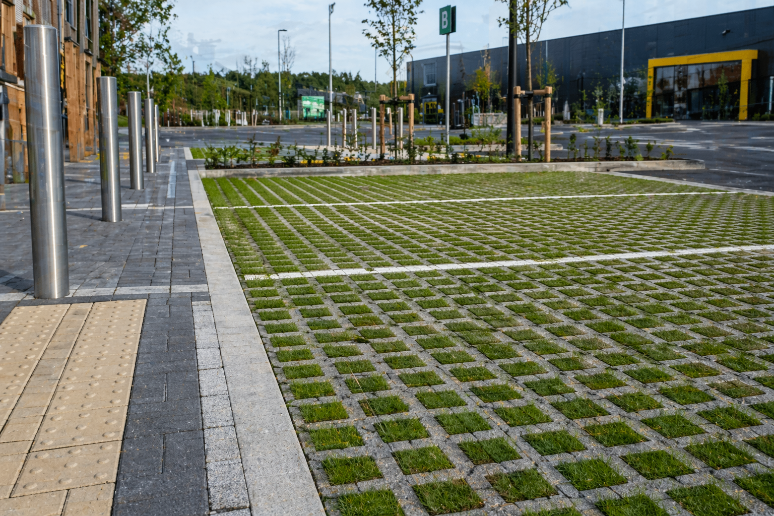 Empty parking lot with grass crete surfacing, metal poles, and a modern commercial building in the background.