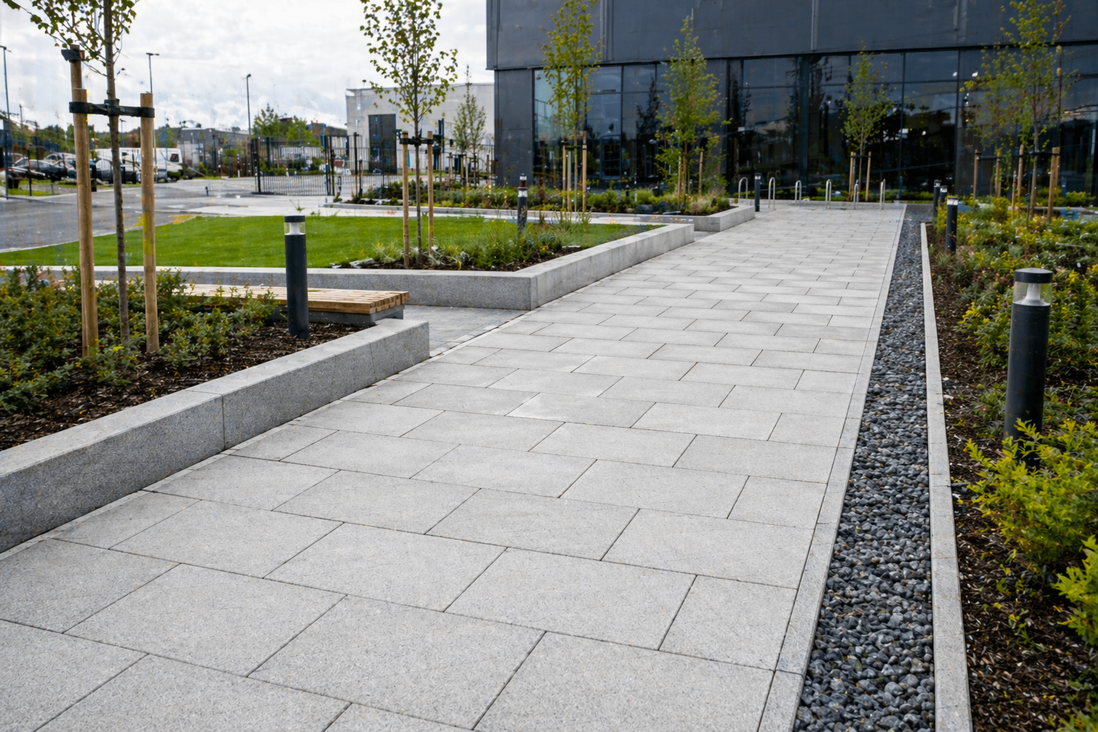 Modern sidewalk with concrete pavers, landscaped areas with trees and shrubs, black bollard lights, and a large glass building in the background.