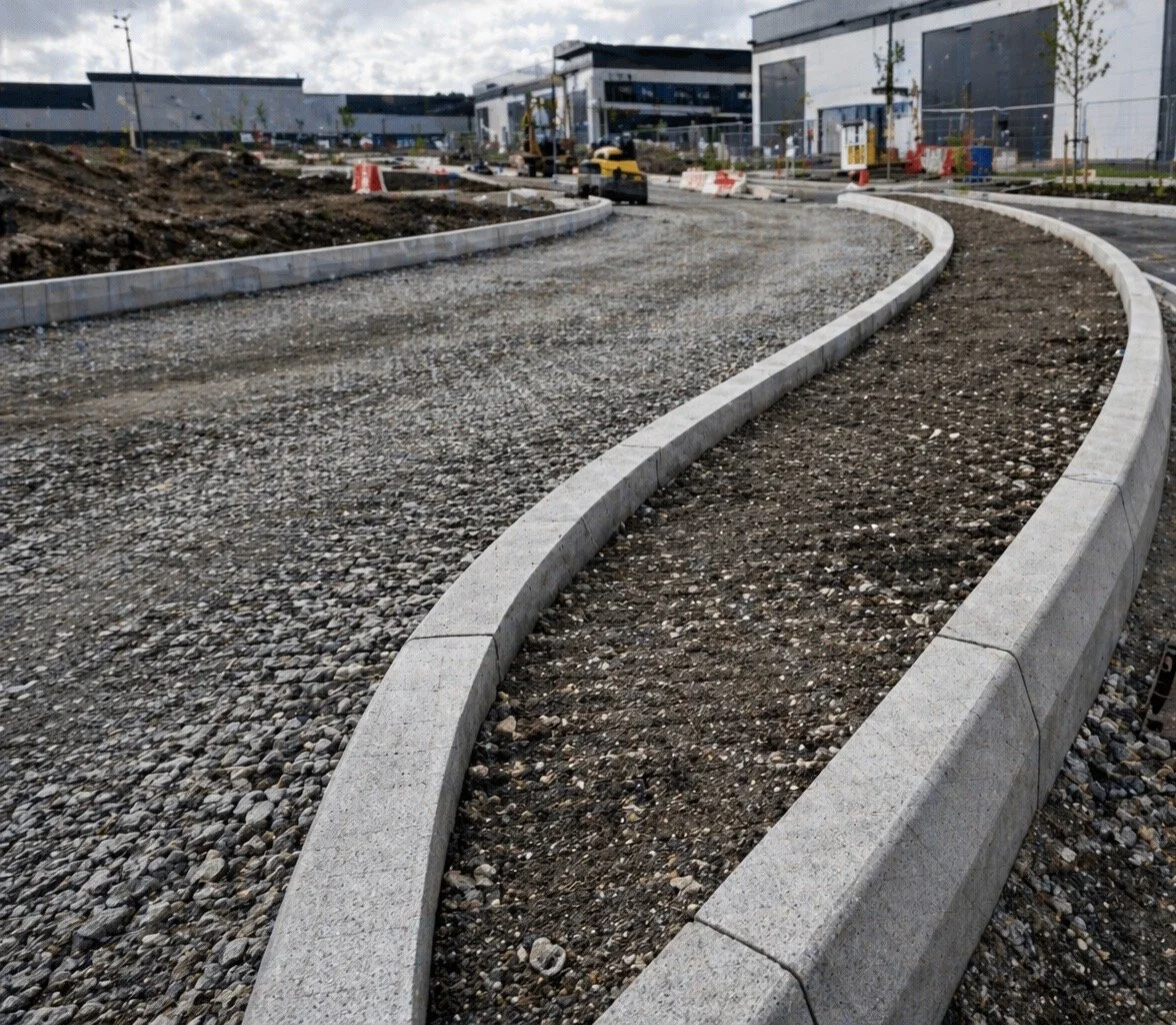 Construction site with curved concrete curb, gravel, and dirt in front of modern glass commercial buildings.