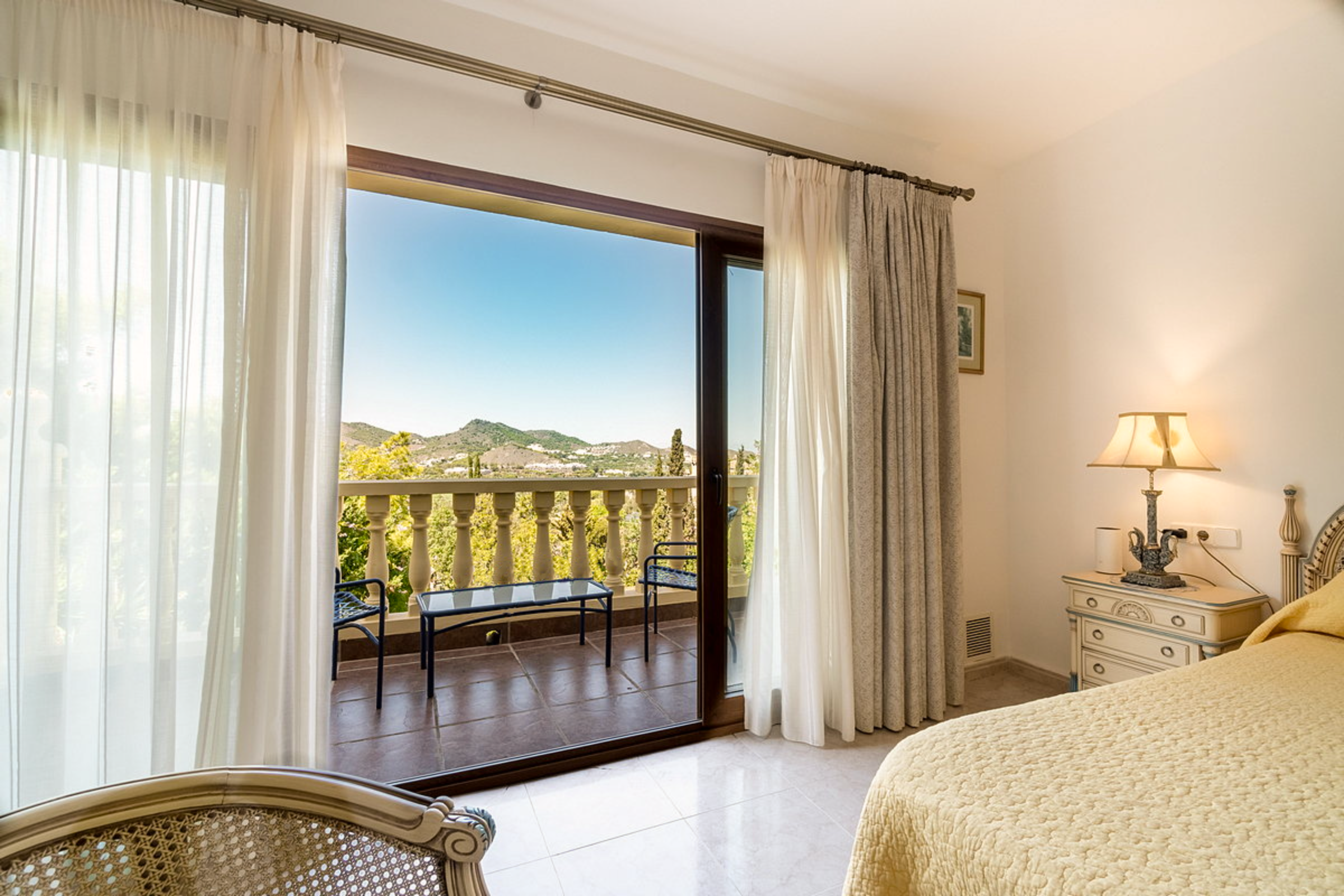 A bedroom with a sliding glass door opening to a balcony with a scenic mountain view. The room has cream-colored walls, a nightstand with a lamp, and a cream quilted bedspread. Sheer white curtains and heavier beige drapes frame the door.