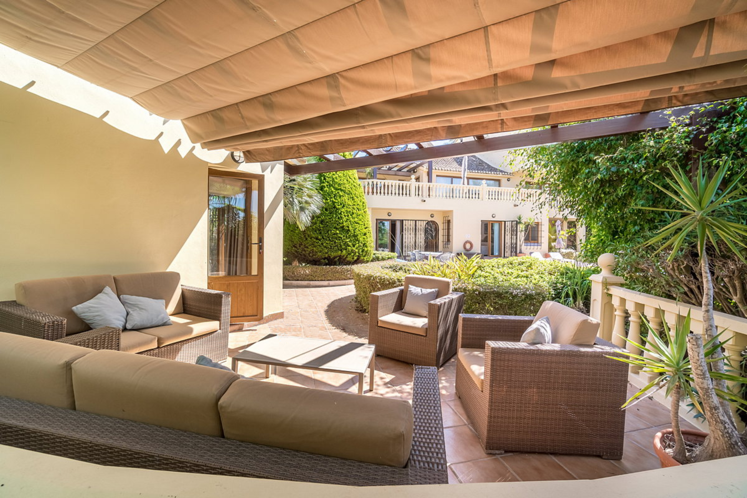 Outdoor patio with wicker and cushioned seating, shaded by a wooden pergola, surrounded by green bushes and trees, with a view of a building with a balcony in the background.