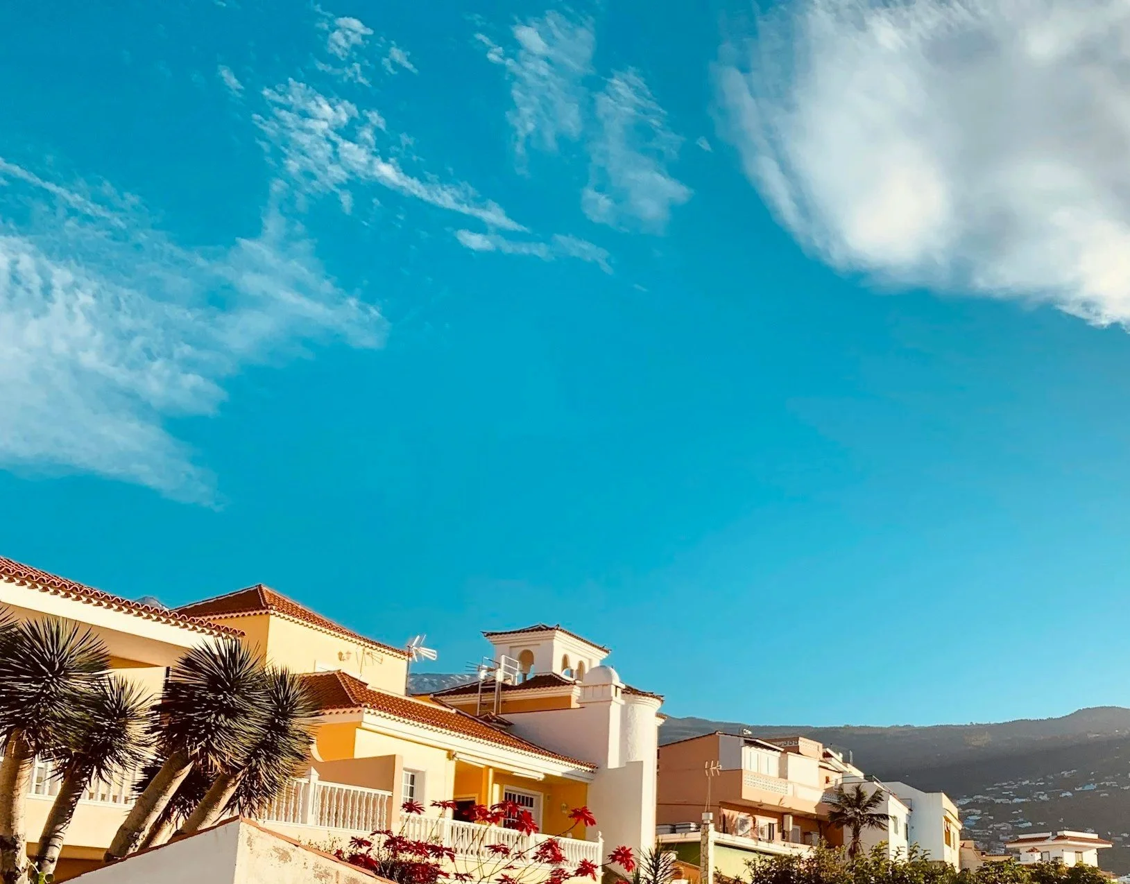 Sunny sky with wispy clouds over colorful Mediterranean-style houses with terracotta roofs and palm trees.