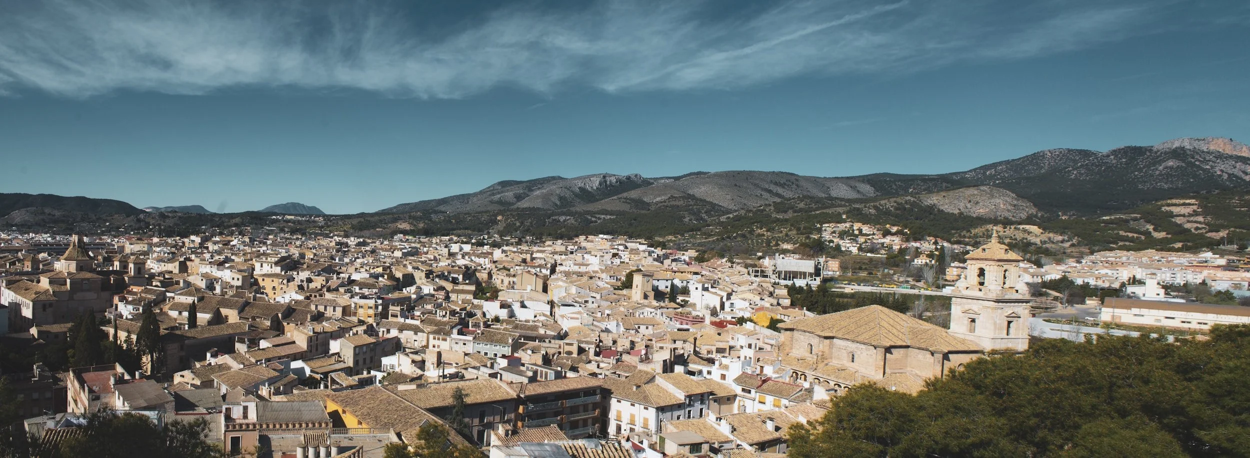 A panoramic view of a town with numerous beige and brown tiled rooftops, surrounded by mountains under a clear blue sky.