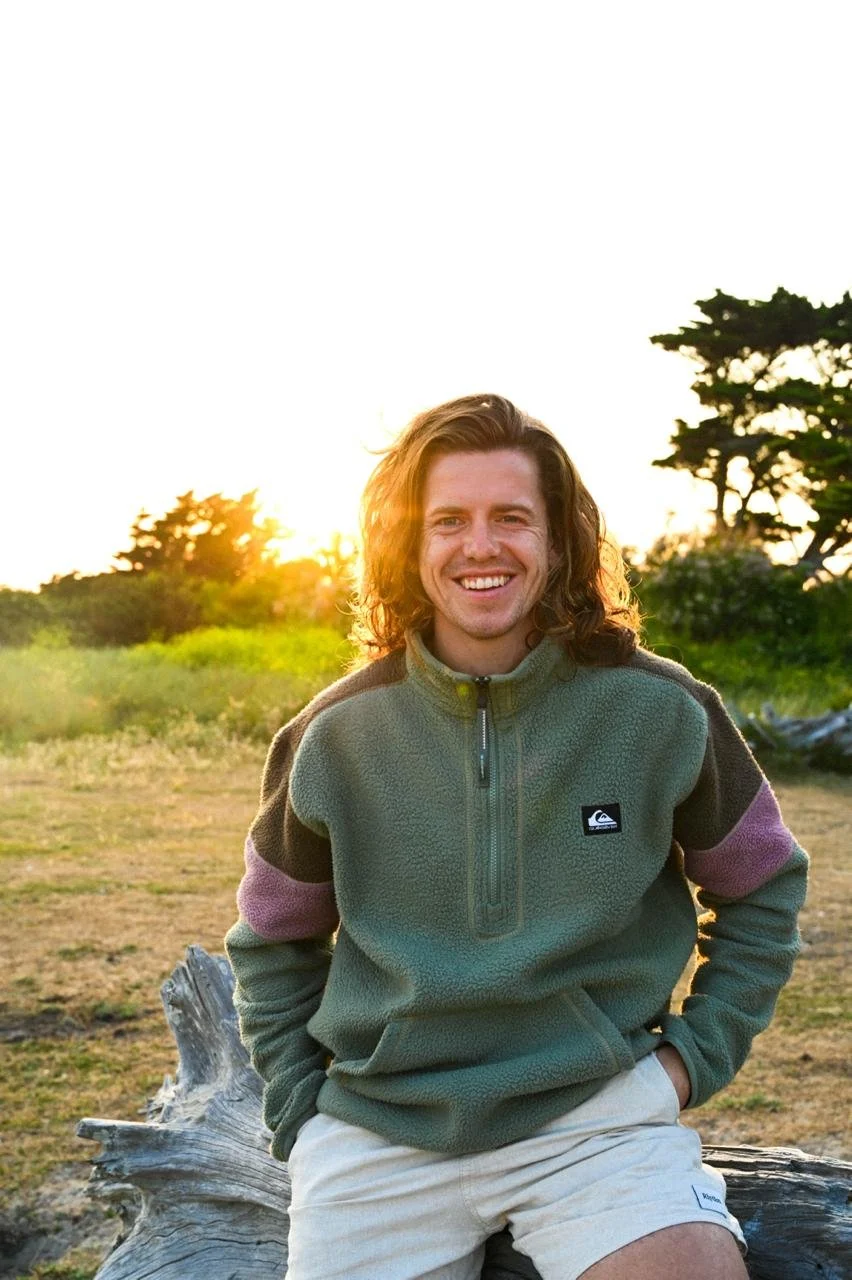 Smiling man sitting outdoors on a tree trunk during sunset with trees and open field in the background.