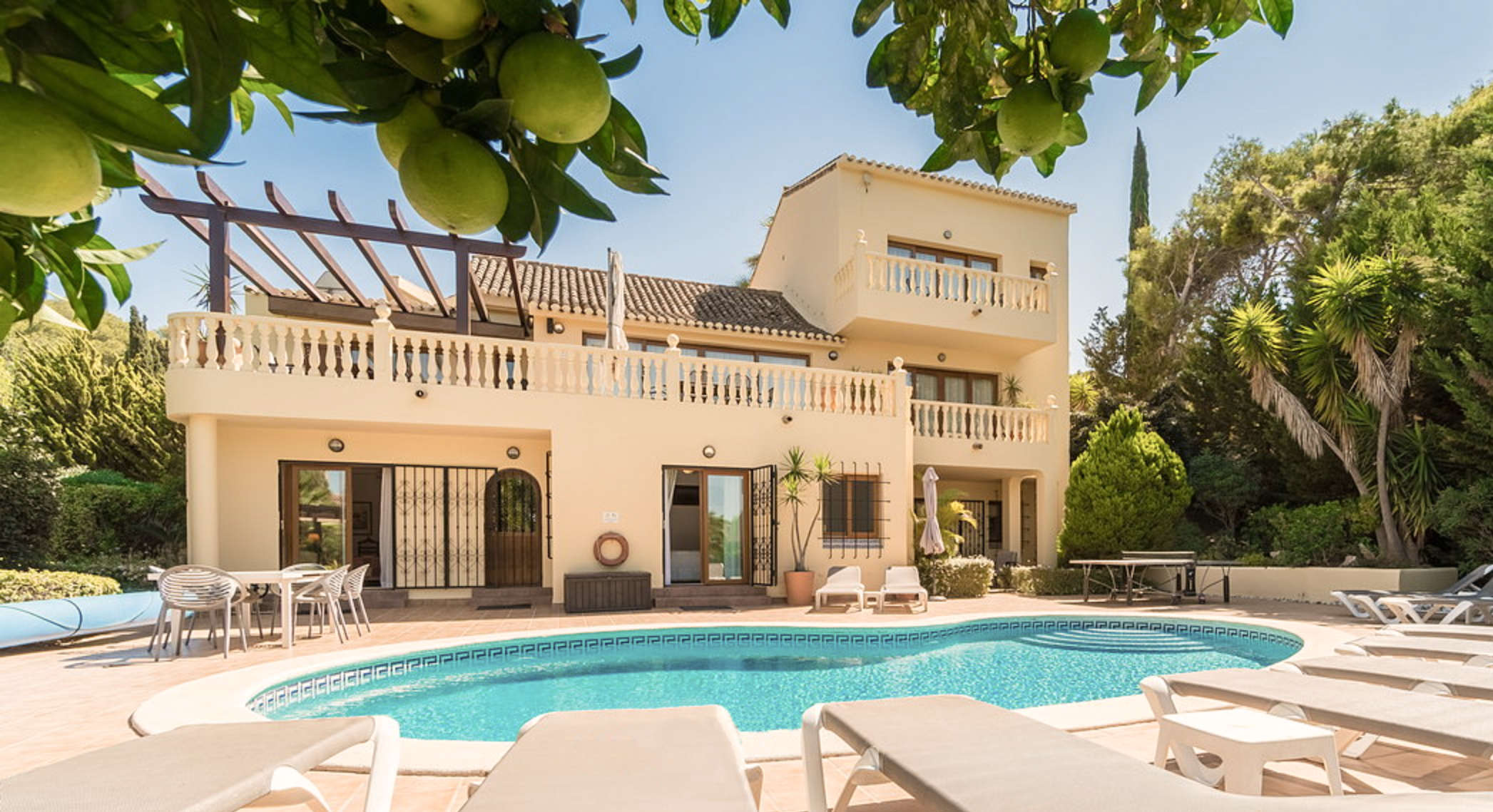 A house with a swimming pool in the backyard, surrounded by trees and lounge chairs, against a clear blue sky.