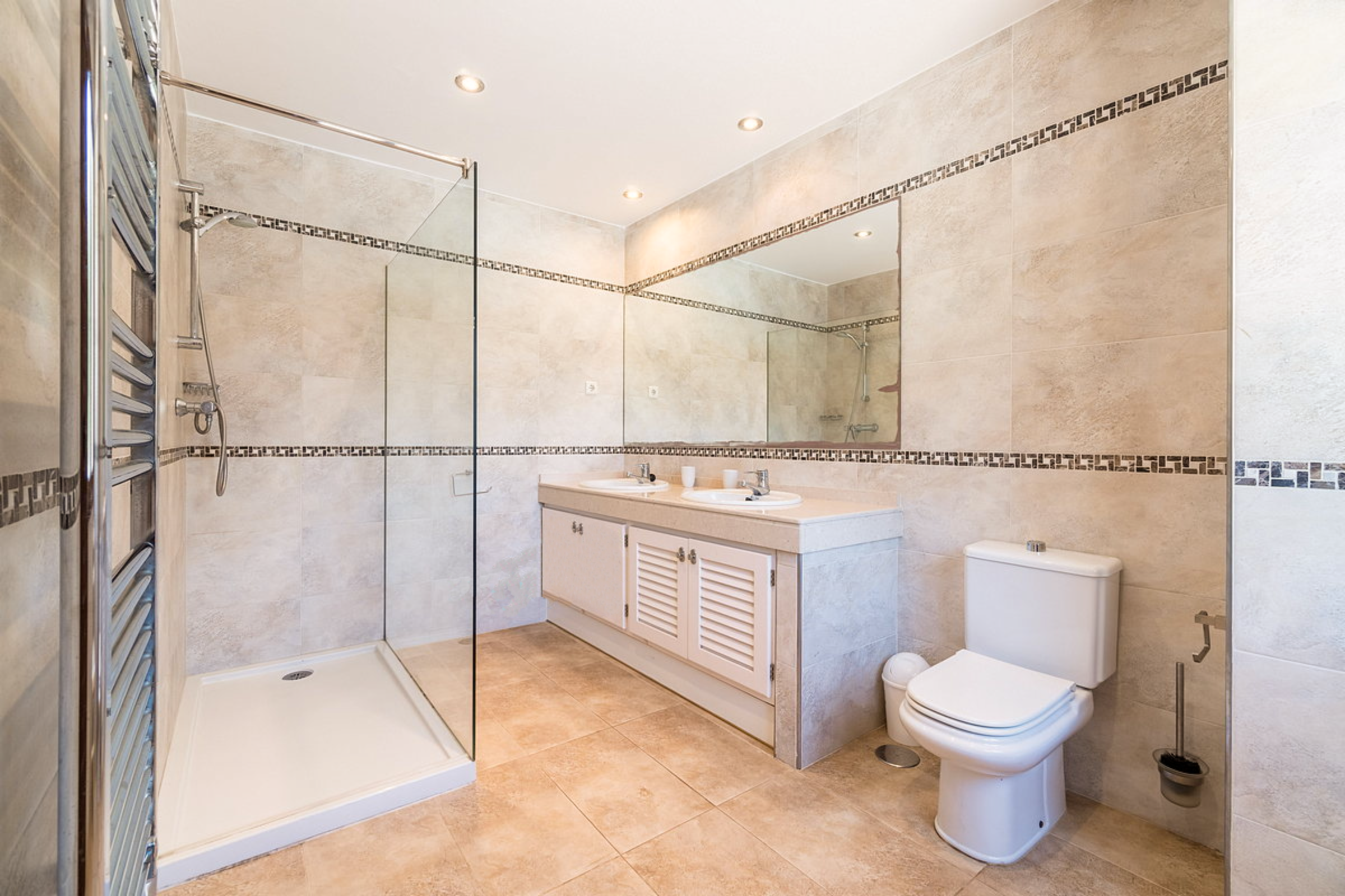 Bathroom with beige tiled walls and floors, including a walk-in shower with a glass divider, dual sinks with mirror, a toilet, and a wall-mounted towel radiator.