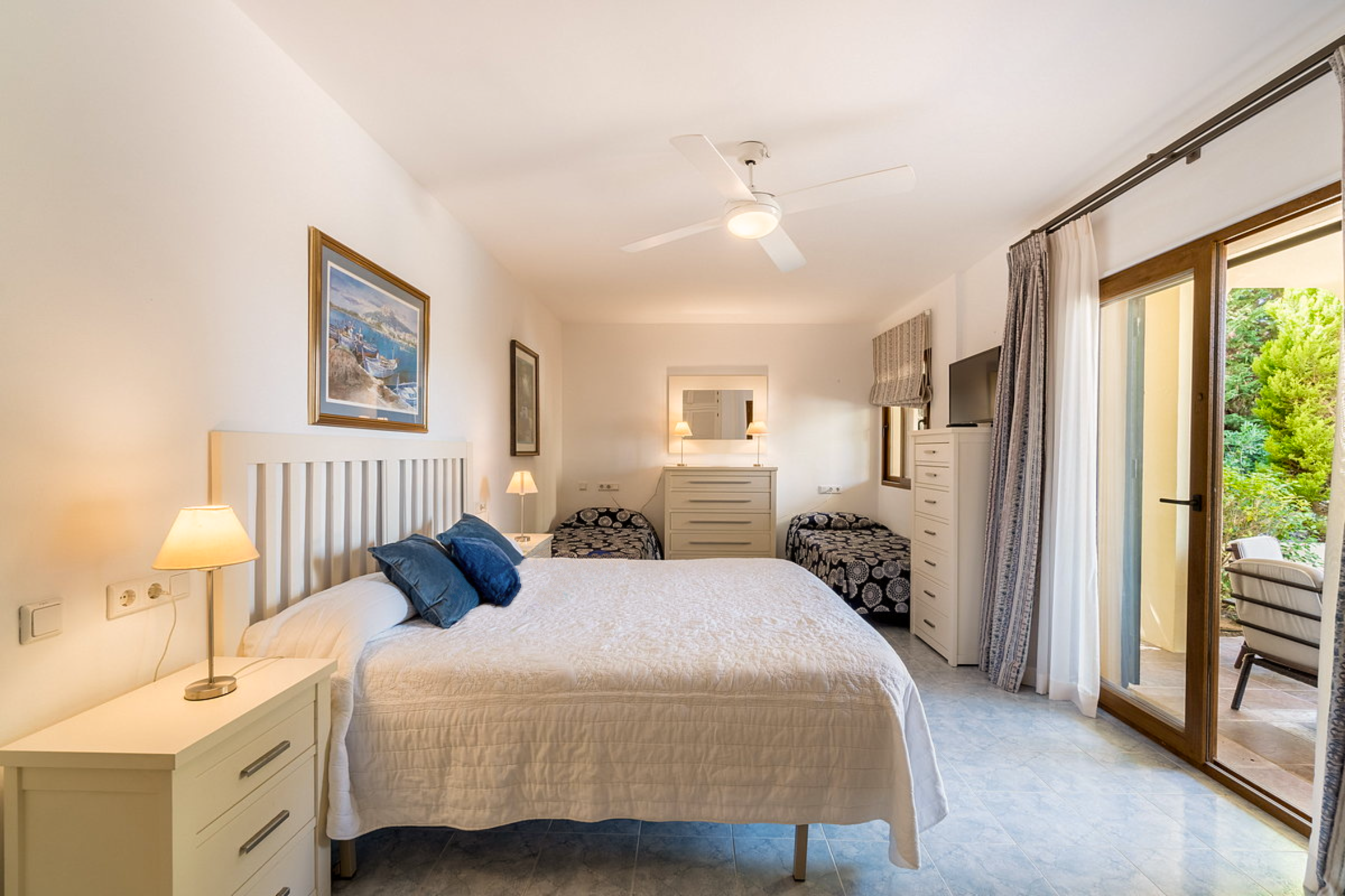 A bedroom with a large bed with white bedding and dark blue pillows. There are two nightstands with lamps on either side of the bed. There are two small patterned chairs at the foot of the bed. A dresser with a mirror and lamps is across the room. There are framed pictures on the walls, and a sliding glass door with curtains leads to a balcony with outdoor furniture and greenery outside.