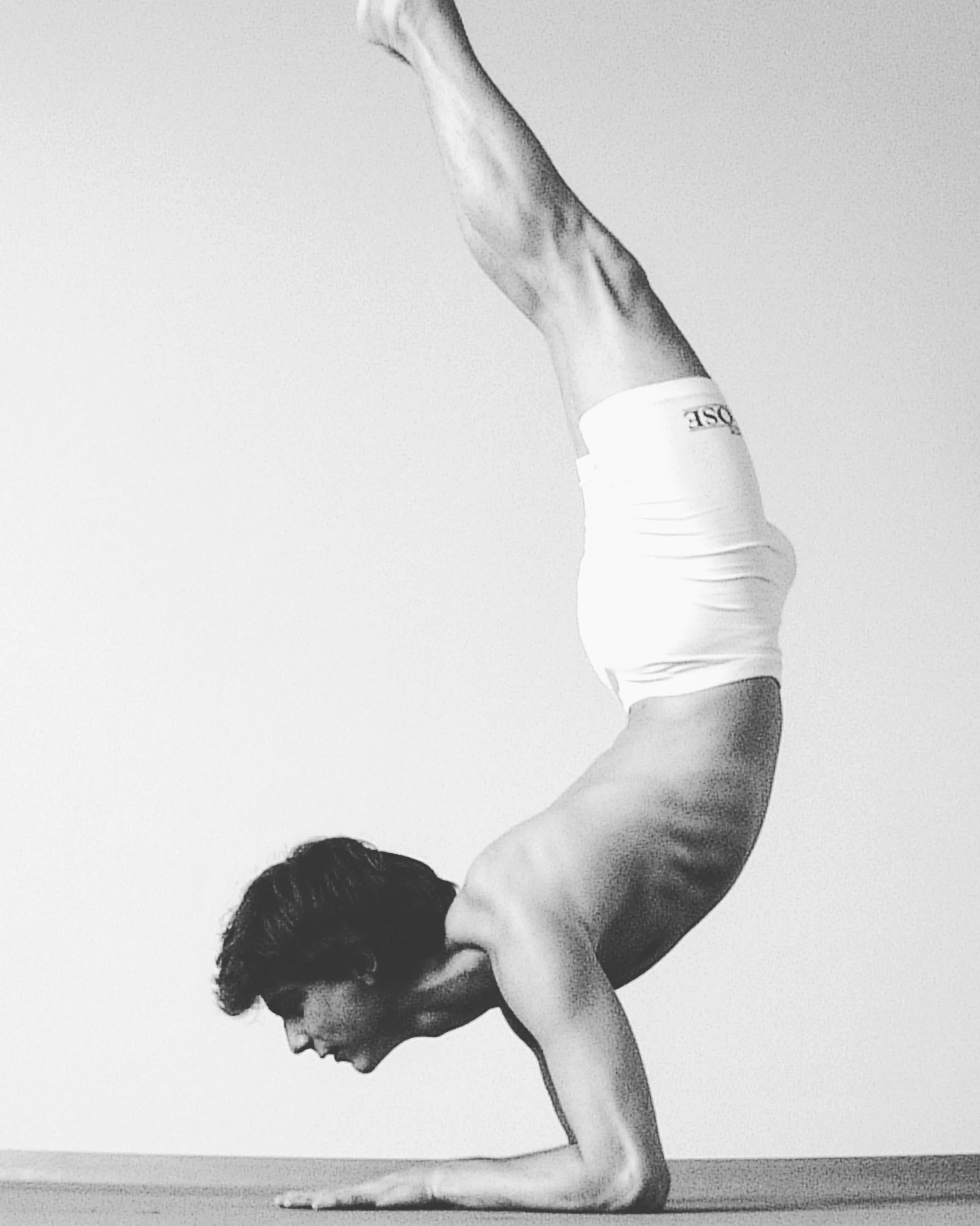 Black and white photo of a woman with dark hair performing a yoga headstand pose, balancing on her head with legs extended upward, against a plain light background.