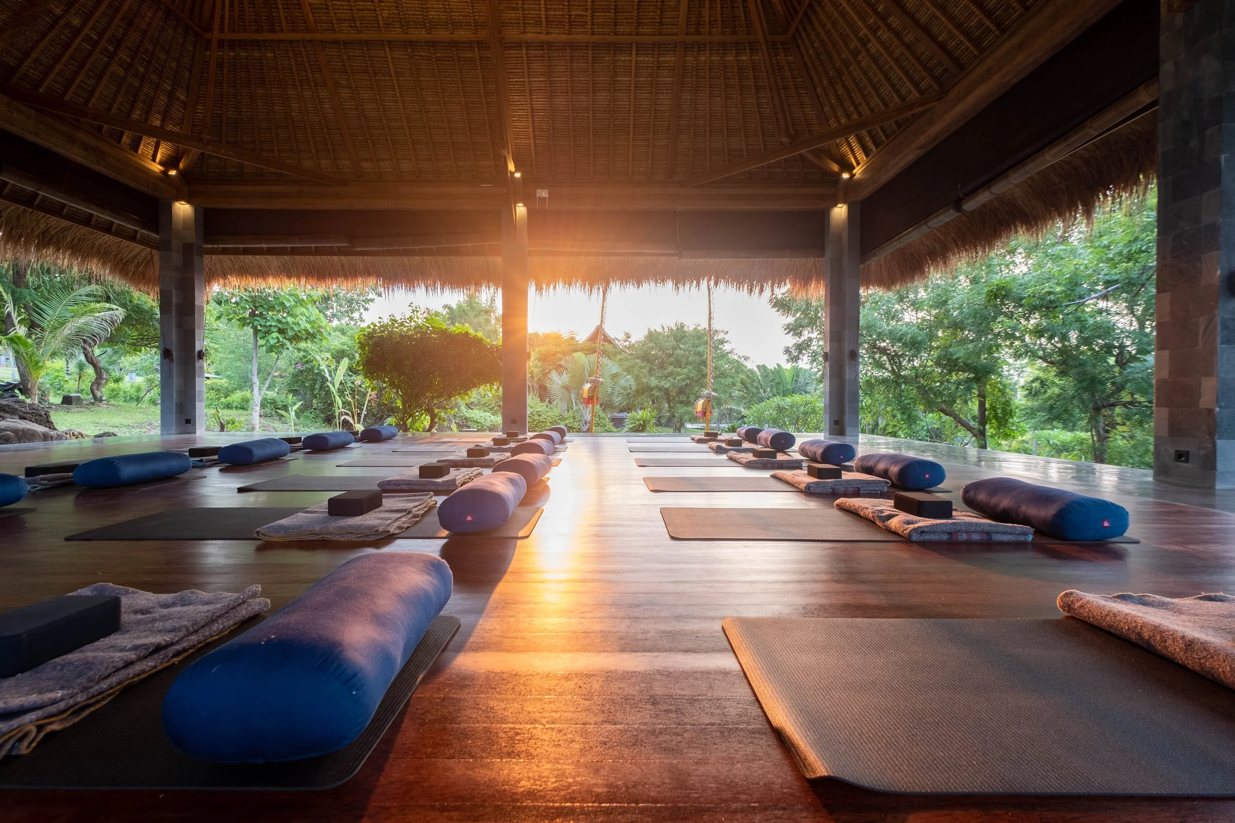 Open-air yoga or meditation pavilion with mats and bolsters arranged on a wooden floor, overlooking lush greenery at sunset.