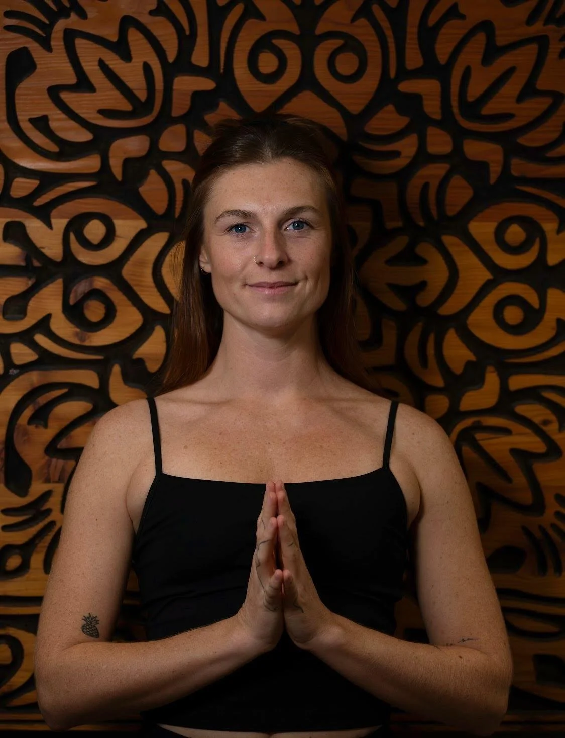 A woman with long brown hair, blue eyes, and freckles standing in front of a decorative wooden wall, wearing a black spaghetti strap top, with her hands pressed together in a prayer position.